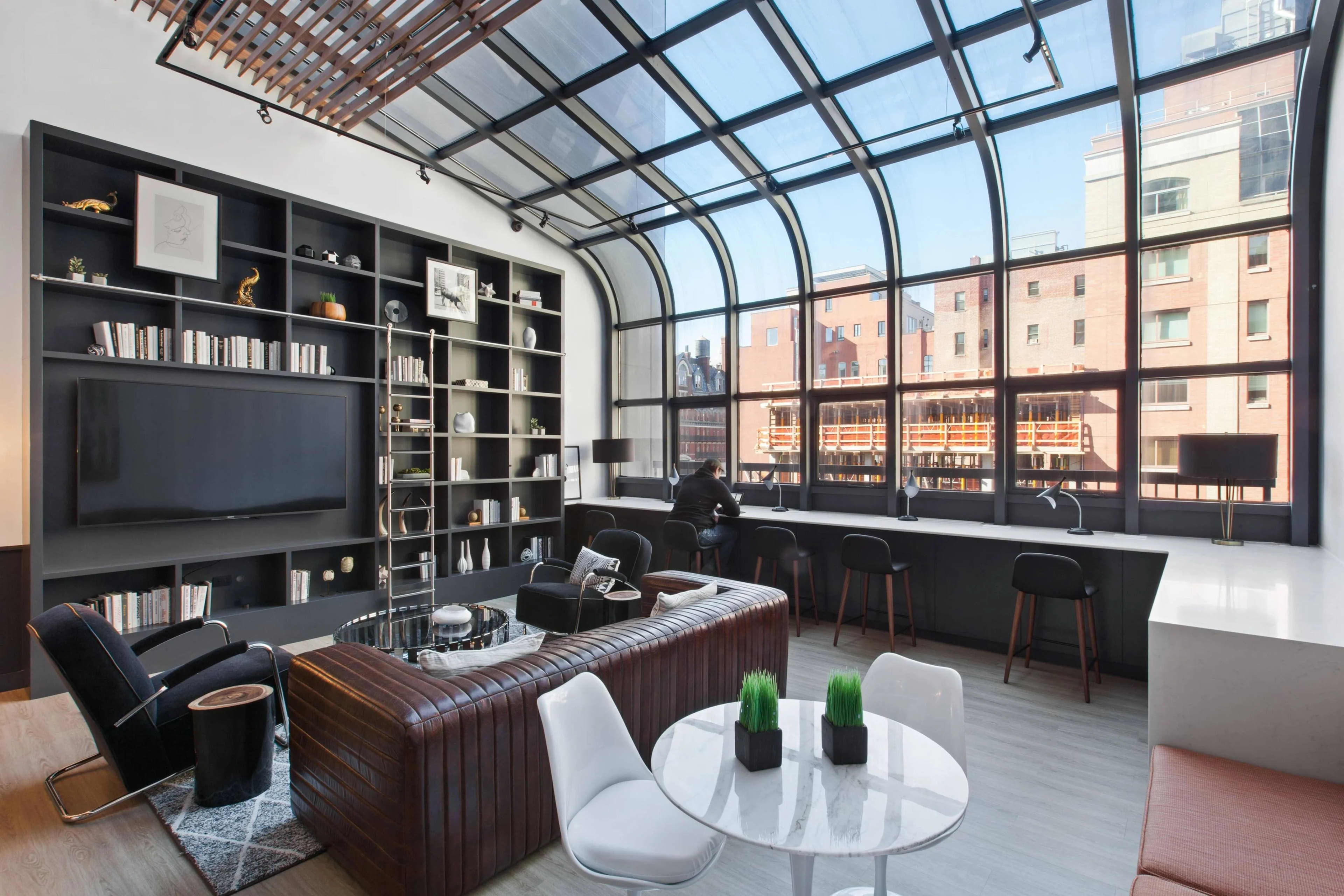 Modern living room with brown leather sectional, white dining table, floor-to-ceiling gridded windows overlooking city buildings, and dark built-in shelving with TV.
