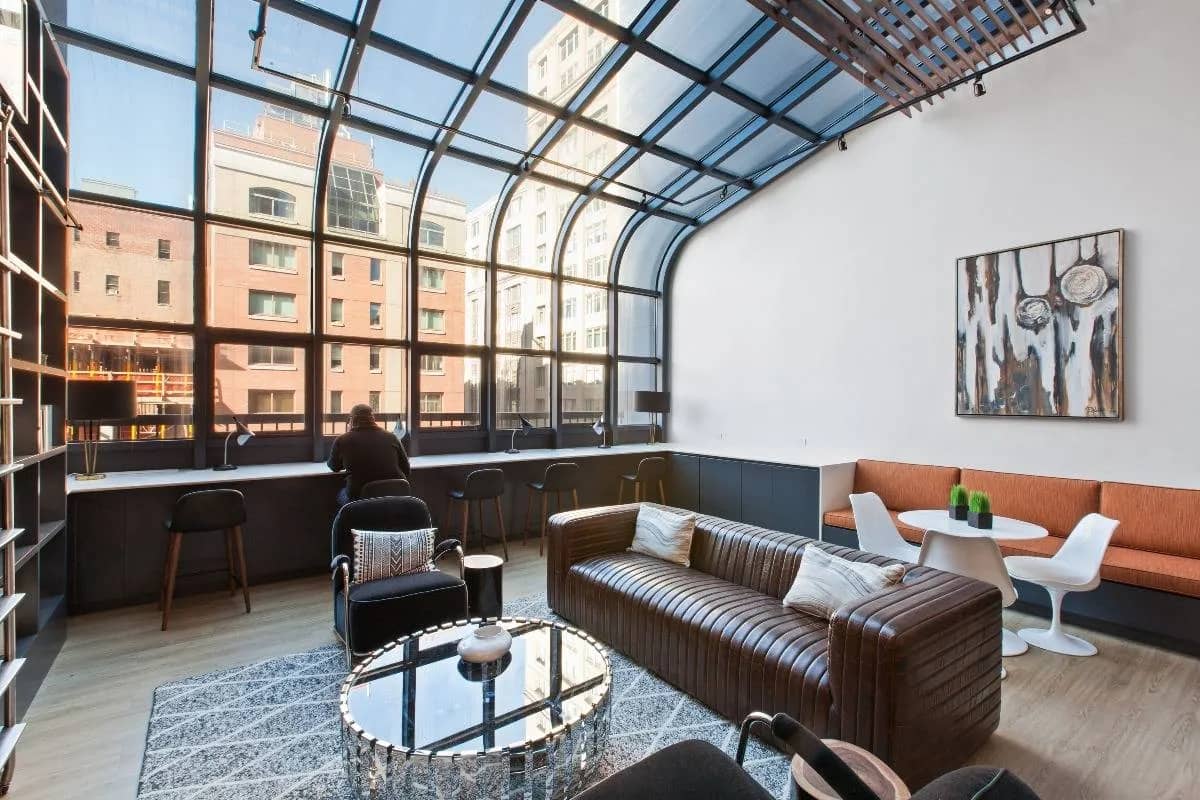 Open-concept loft living room with brown leather sofa, glass-top coffee table, floor-to-ceiling windows overlooking city buildings, and contemporary artwork.