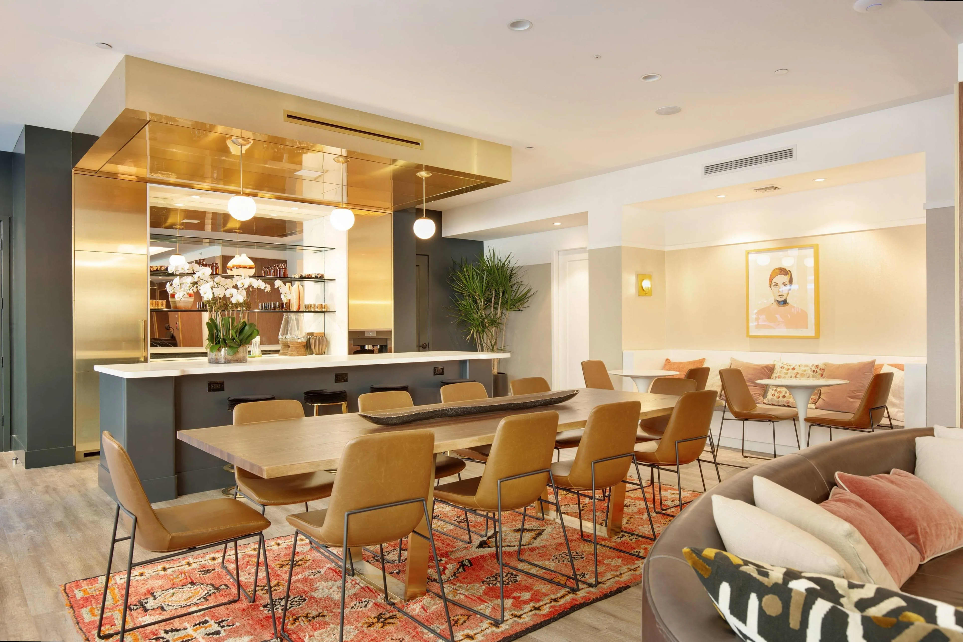 Open-concept common room with dining table, tan chairs, illuminated bar with shelving, and lounge seating with coral accents.