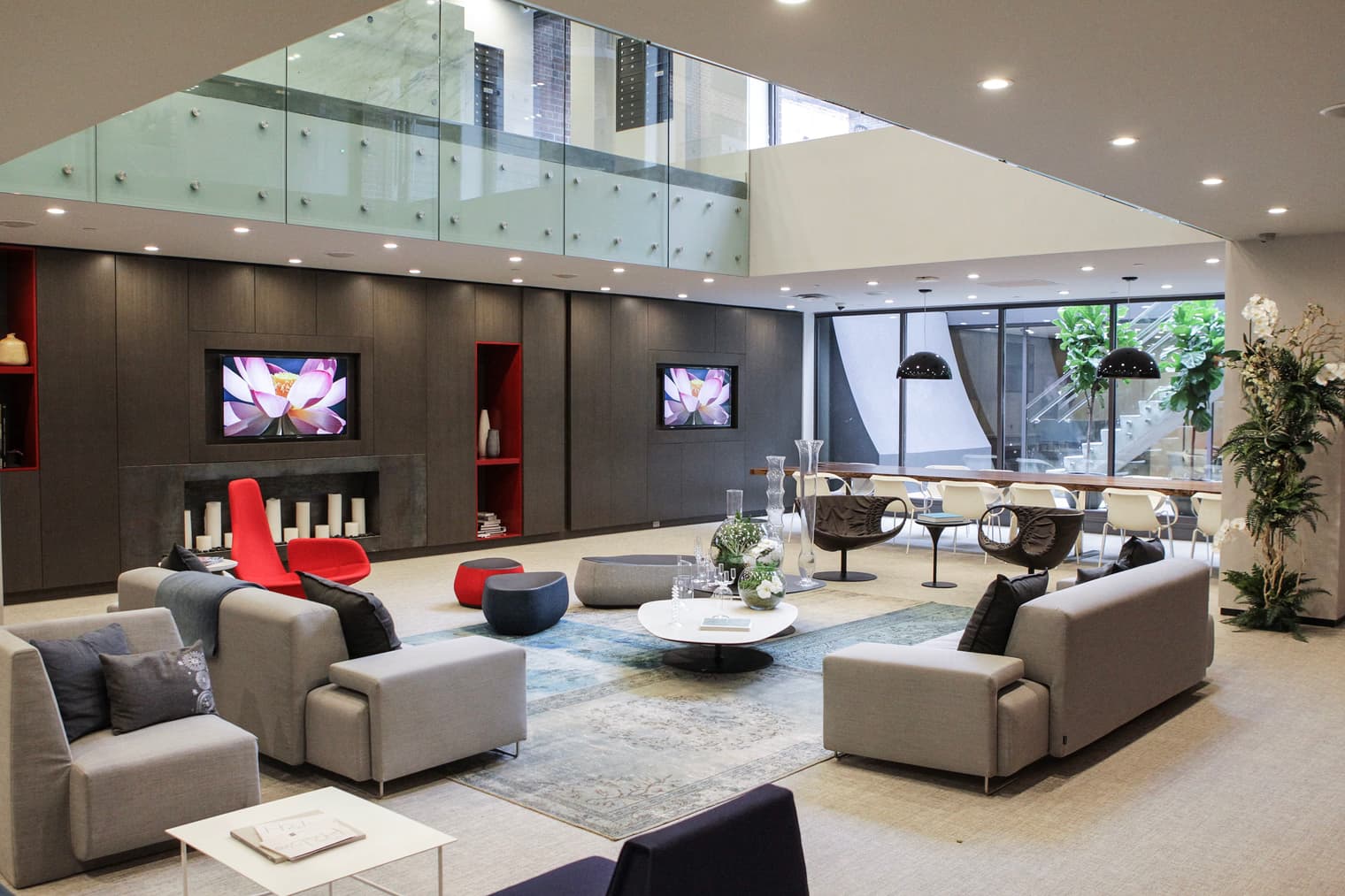 Open-plan lounge with beige sectional sofas, red accent chair, dark wood wall with mounted TV, glass mezzanine above, and large windows with city views.