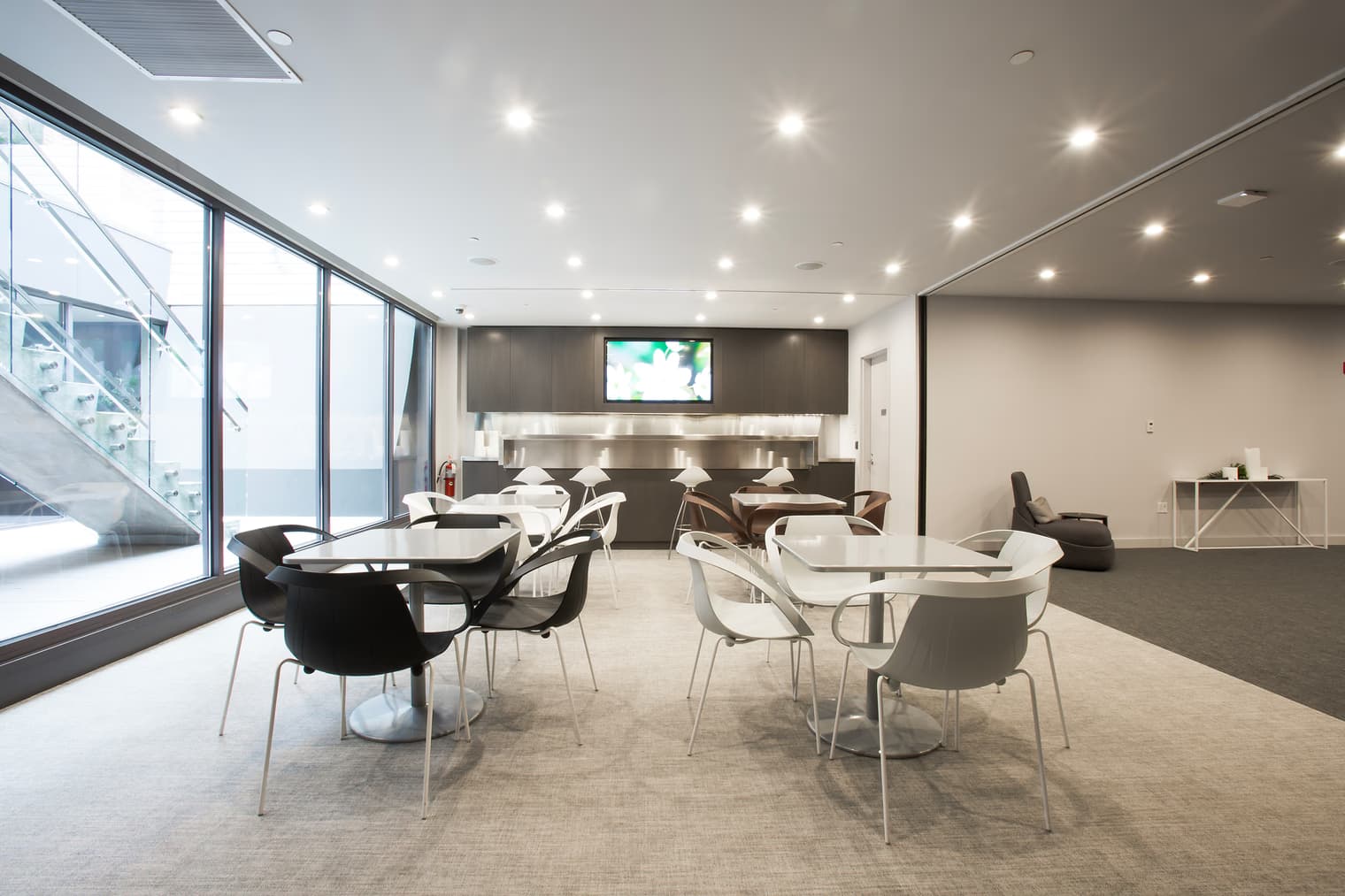 Open common room with round dining tables, black and white chairs, floor-to-ceiling windows, recessed lighting, and kitchen area with wall-mounted TV.