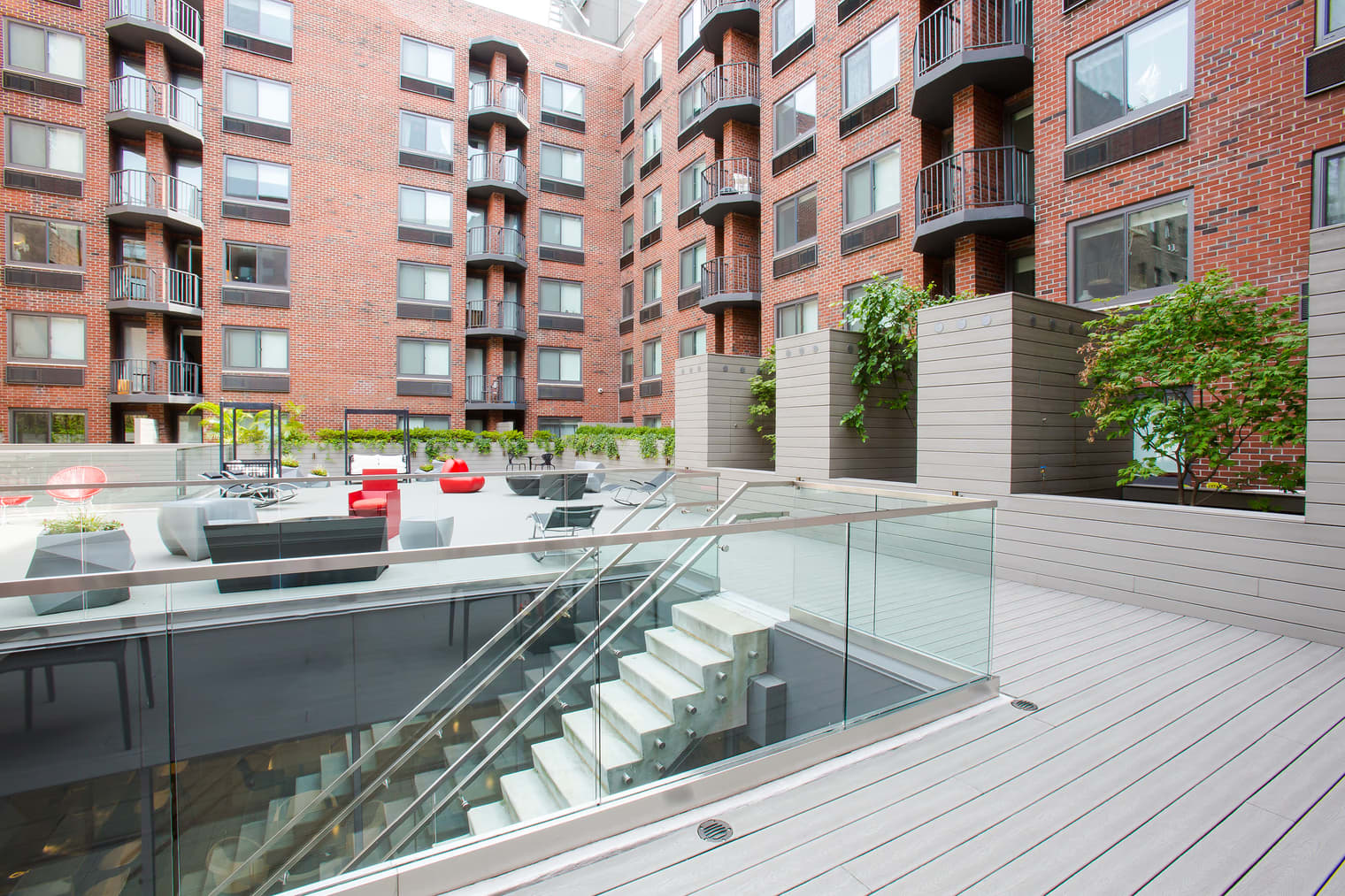 Contemporary courtyard with composite deck, glass-railed staircase, red lounge seating, and ivy-covered planters framed by multi-story brick apartment buildings.