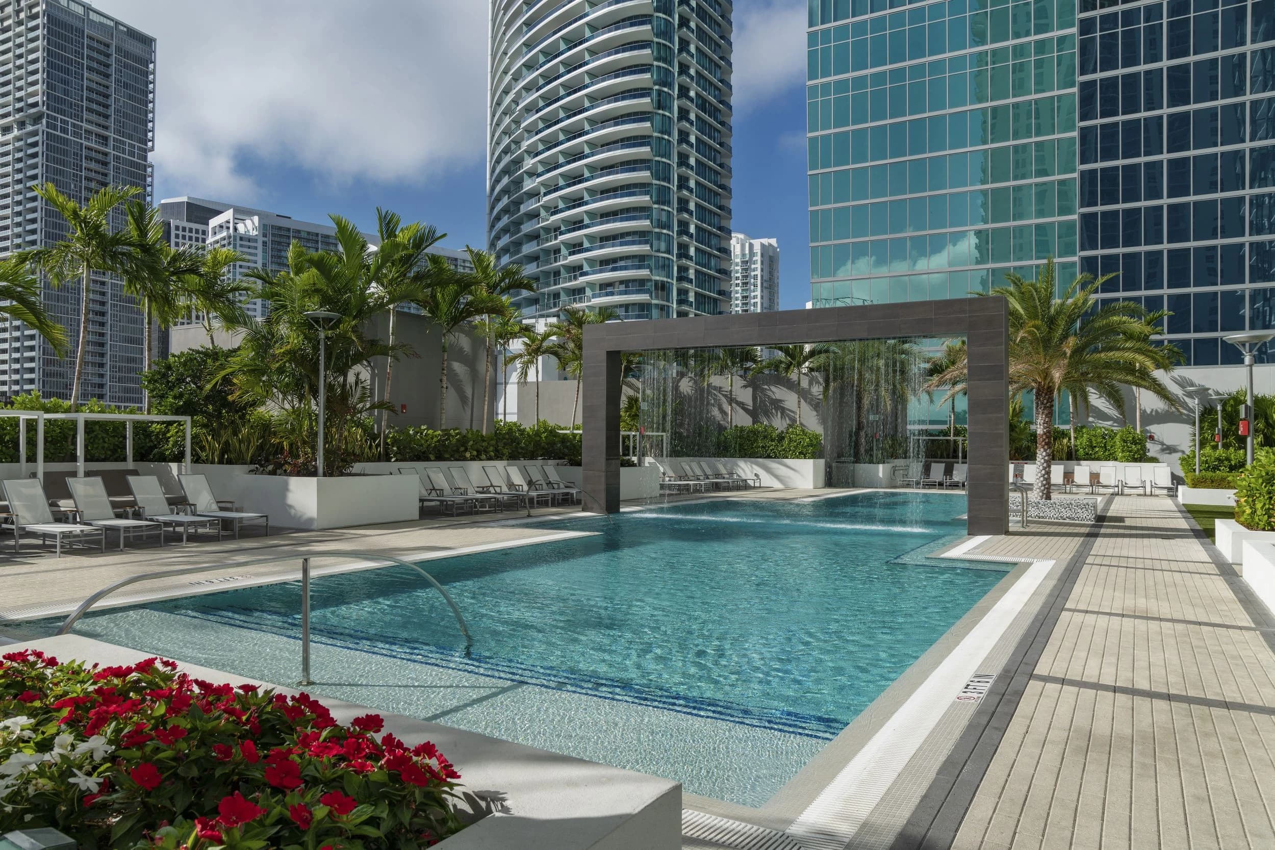Resort pool with clear turquoise water, gray lounge chairs, red flowers, palm trees, modern pergola, and Miami high-rise buildings in background.