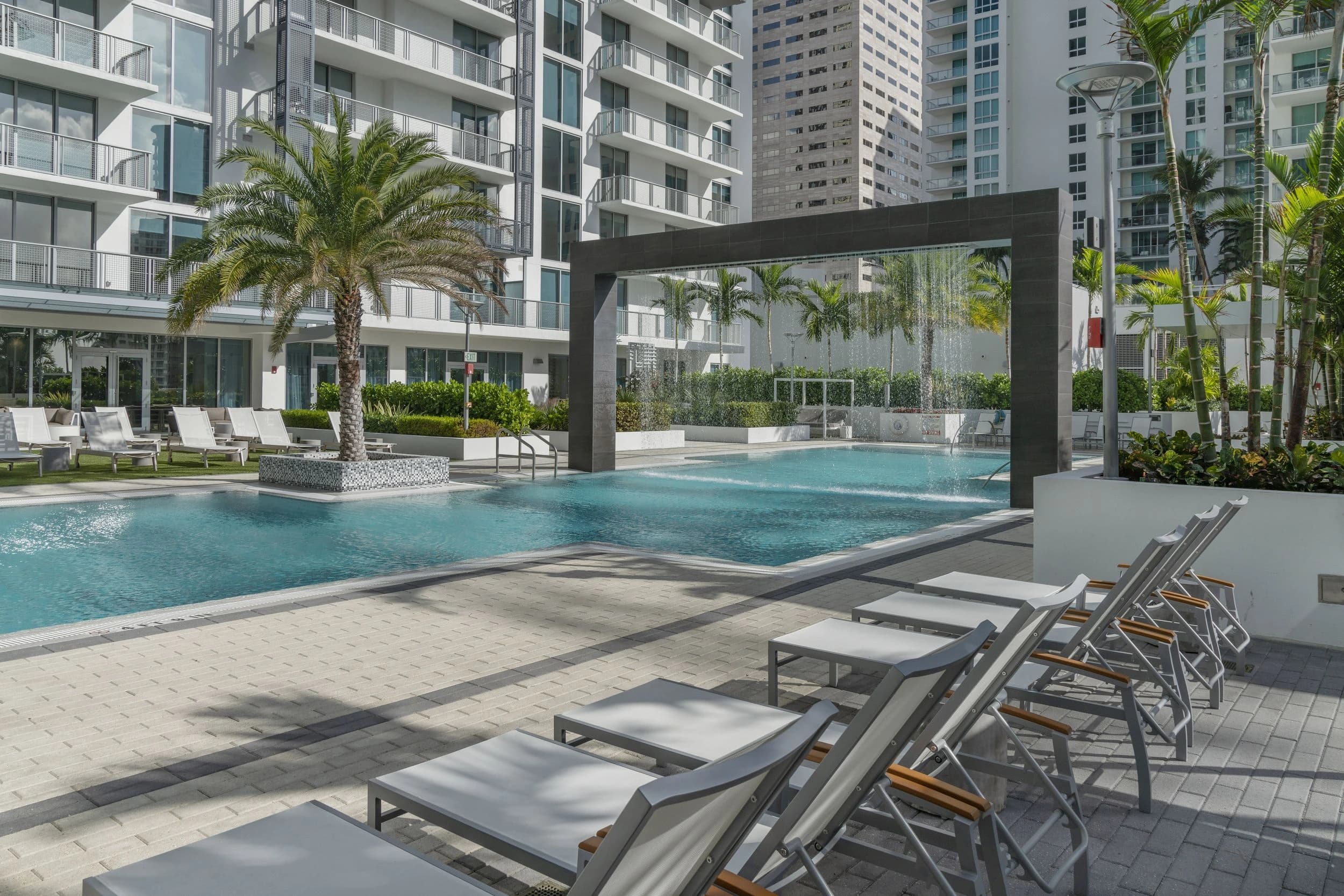 Resort pool with turquoise water, lounge chairs in foreground, black pergola overhead, palm trees, and multi-story residential towers surrounding the deck.