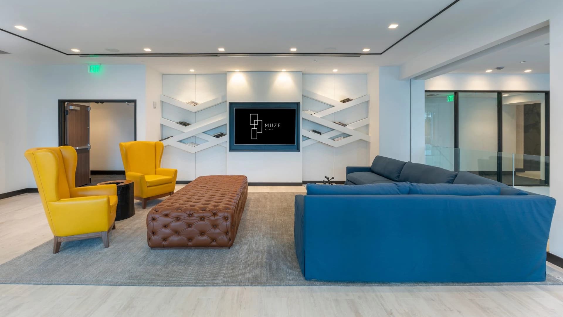 Common lounge with blue sectional sofa, yellow accent chairs, brown tufted ottoman, geometric white shelving, and recessed ceiling lights.