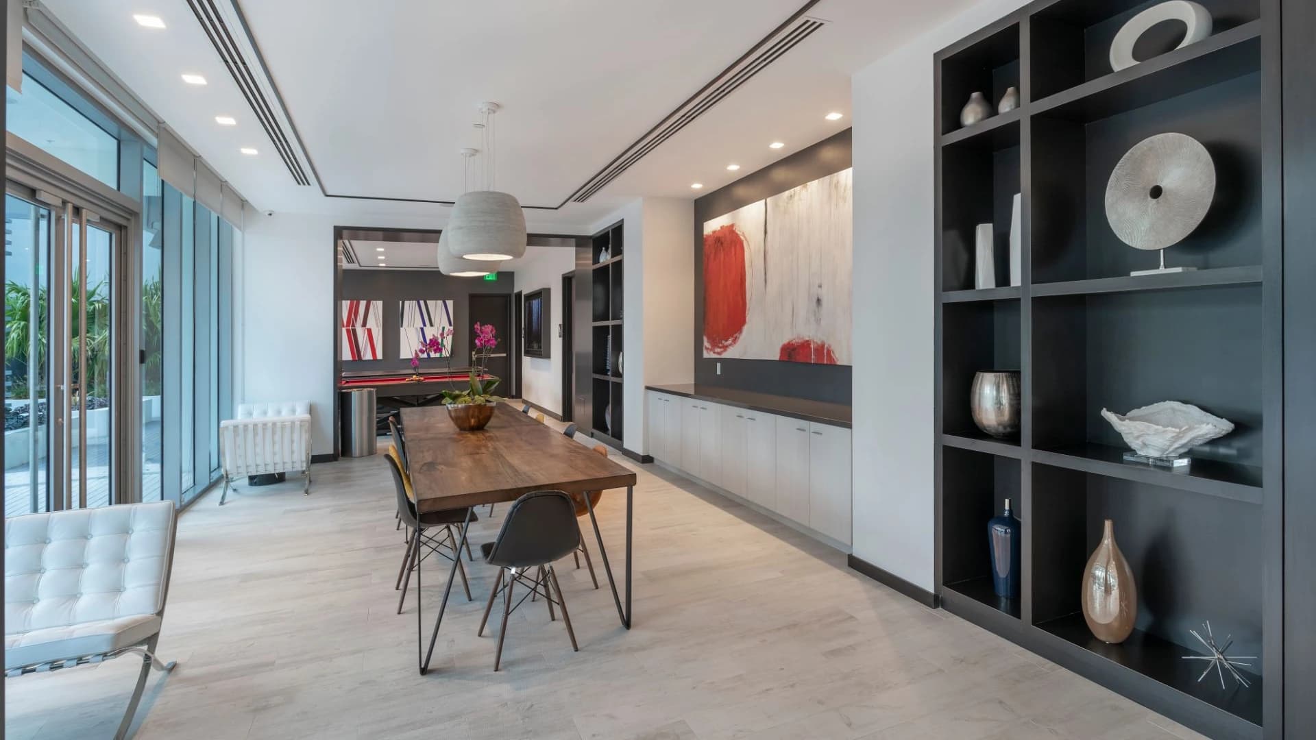 Open-concept living and dining room with wood dining table, black shelving unit displaying decorative objects, pendant lighting, and floor-to-ceiling windows.