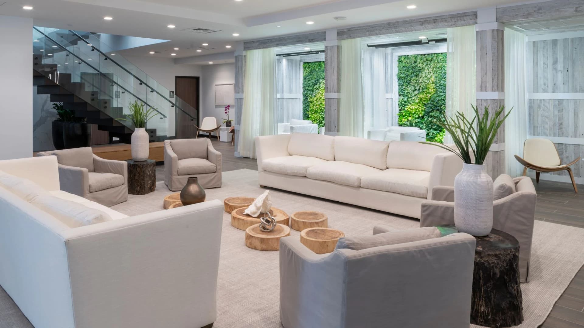 Open living room with cream sofas, wooden accent tables, potted plants, and glass staircase; large windows overlook green outdoor space.
