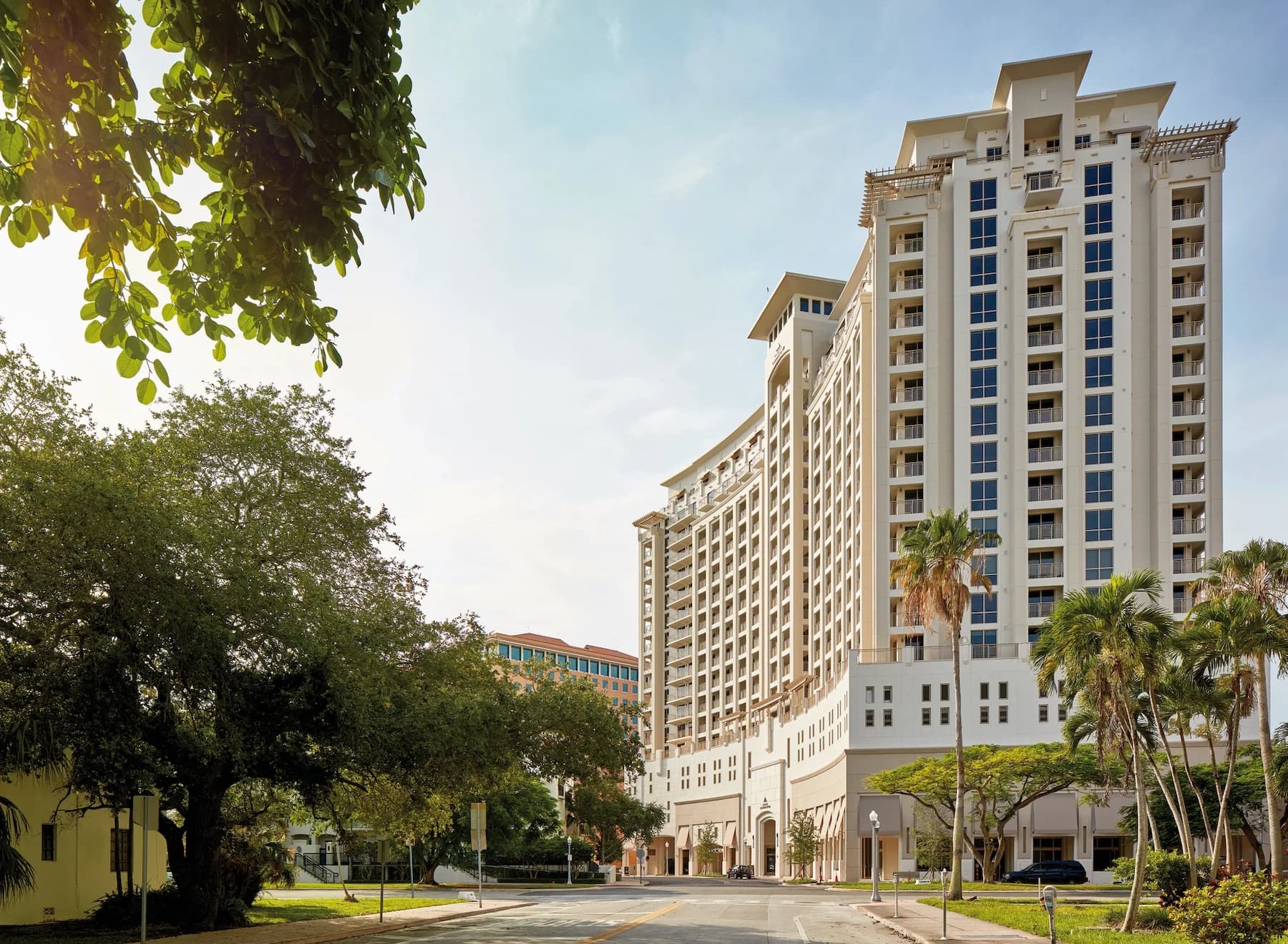 Multi-story residential building with cream and white facade, blue-trimmed windows, and palm trees in foreground under clear sky.