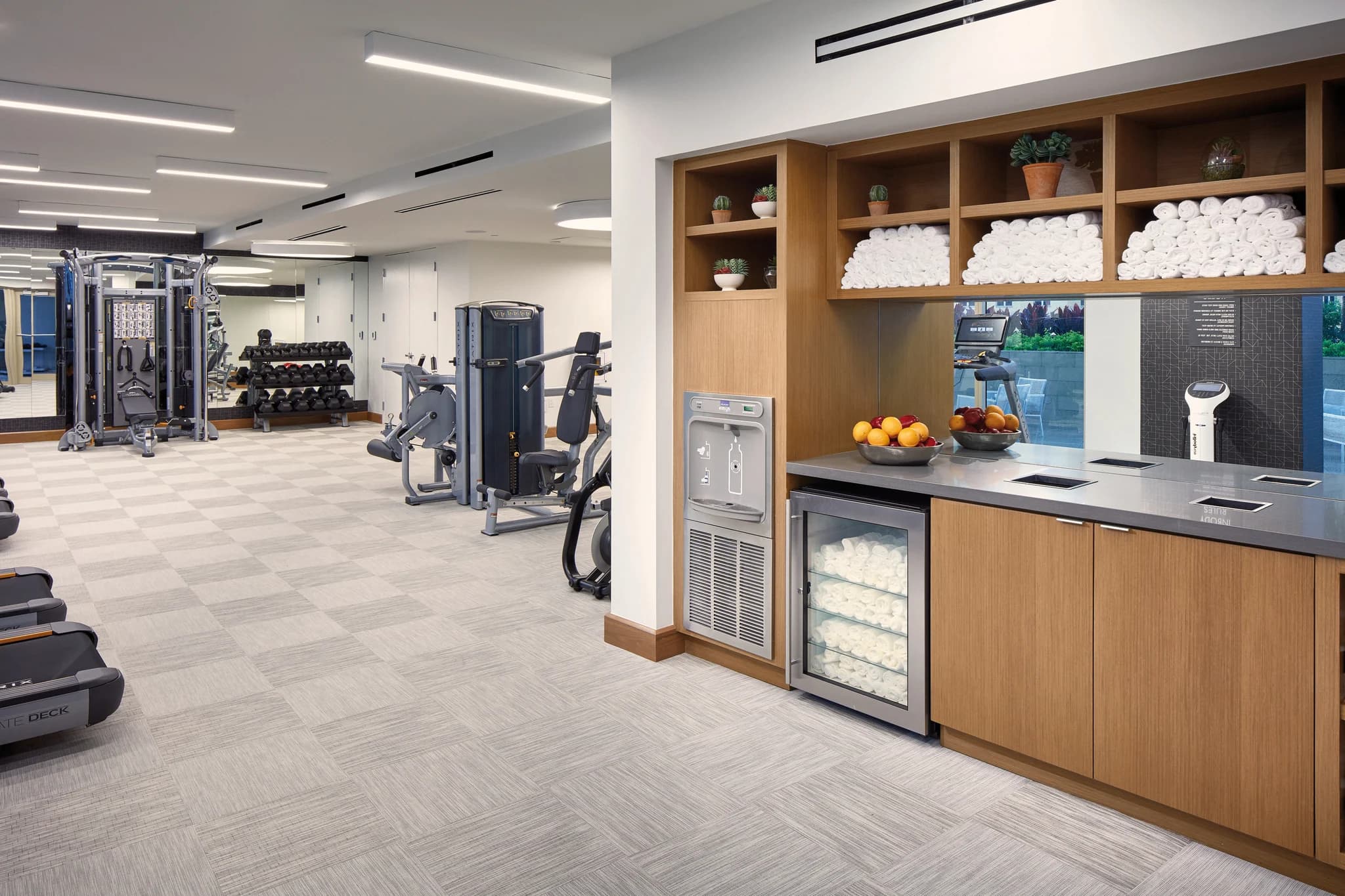 Fitness center with exercise machines, wood cabinetry with white towels, water cooler, and fresh fruit on counter.