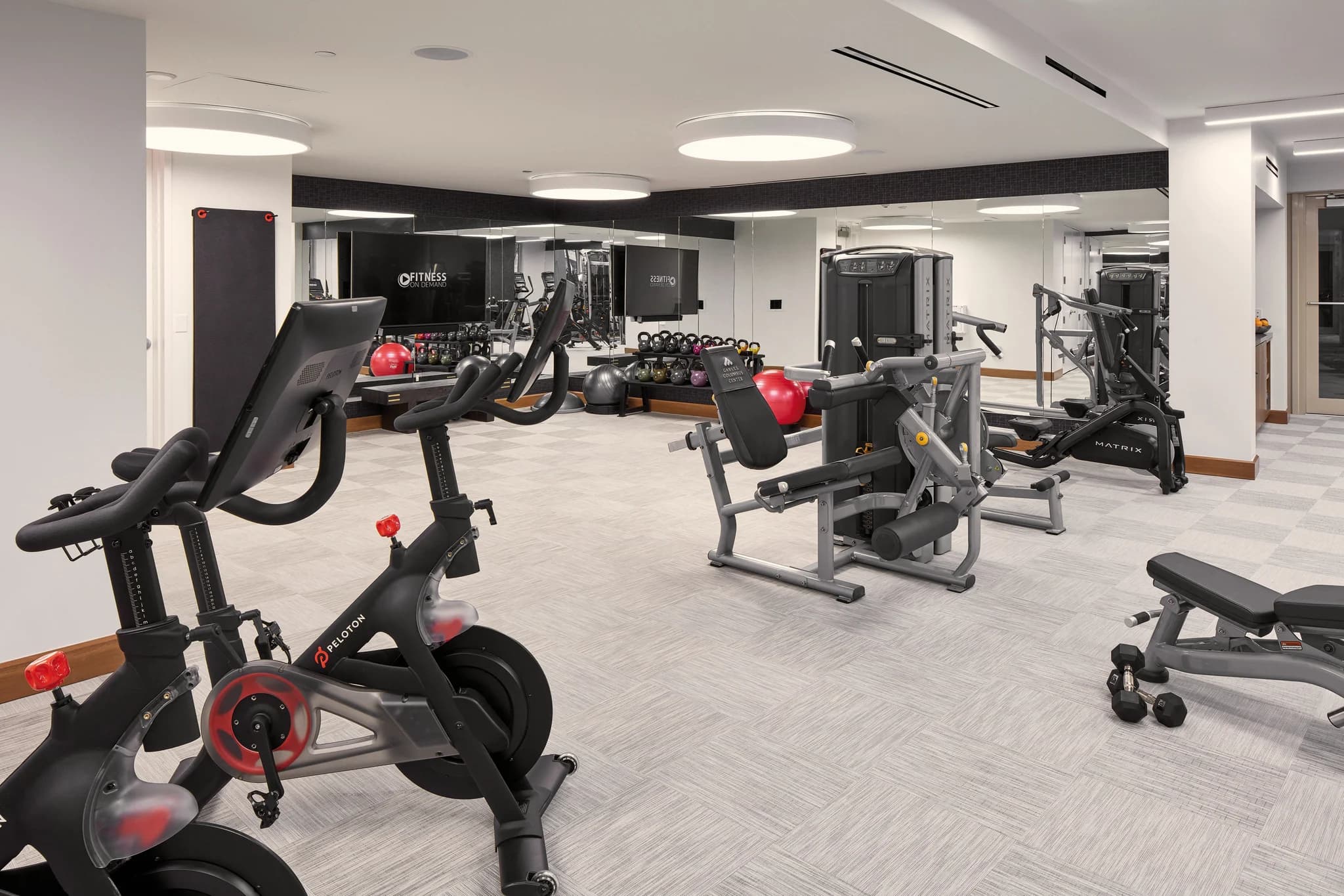 Modern fitness center with stationary bikes in foreground, weight machines, dumbbells, and cardio equipment throughout bright white room with recessed lighting.