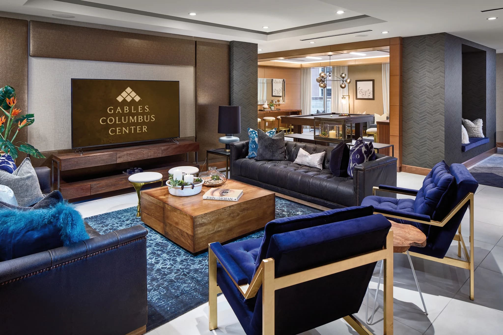 Resident lounge with black leather sofas, blue accent chairs, wooden coffee table, flat-screen TV, and open kitchen bar area.