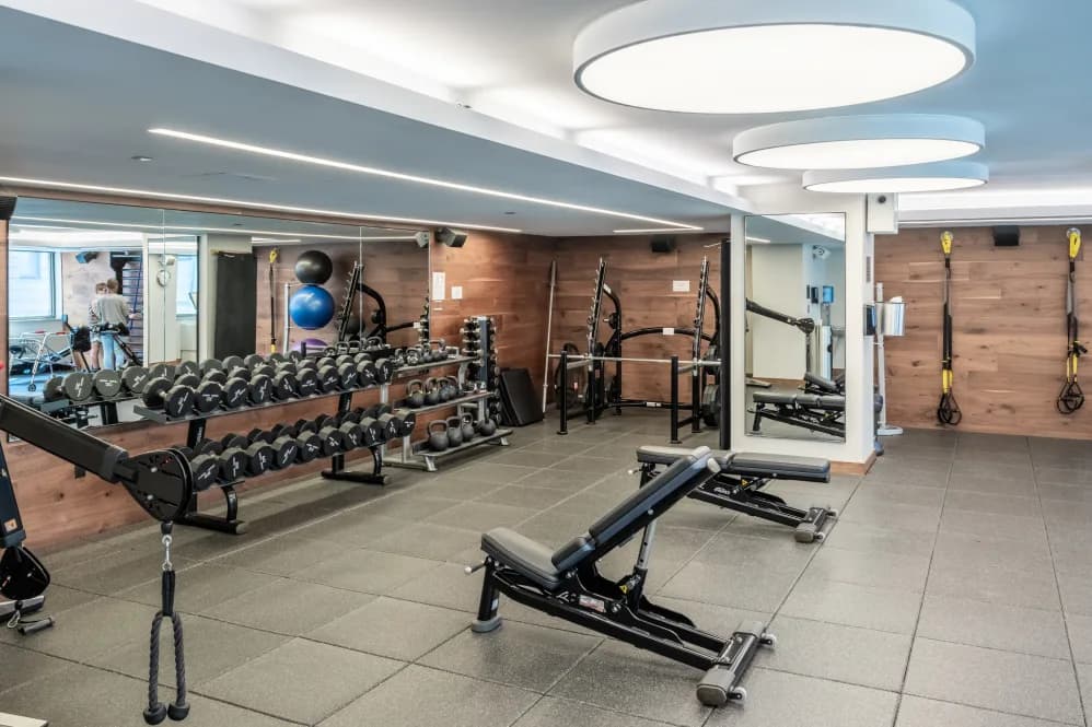 Modern fitness center with rows of dumbbells, exercise bikes, weight benches, and suspension training straps on wood-paneled walls under bright ceiling lights.