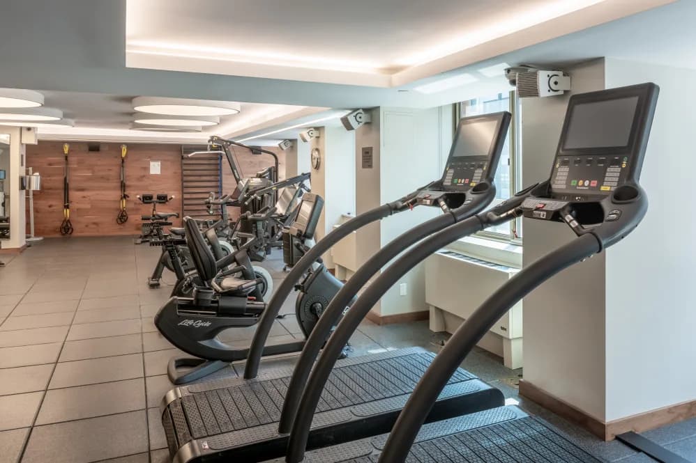 Fitness center with elliptical machines in foreground, stationary bikes, weight rack, and ceiling-mounted screens.