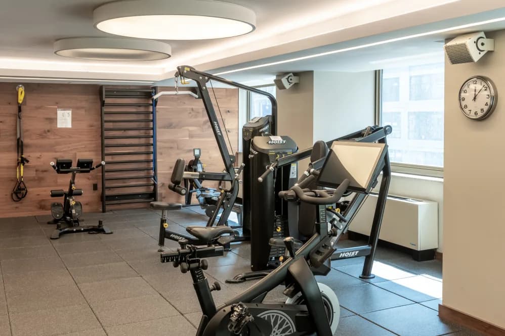 Modern fitness room with stationary bikes, exercise machines, wood accent wall, recessed ceiling lights, and large windows.