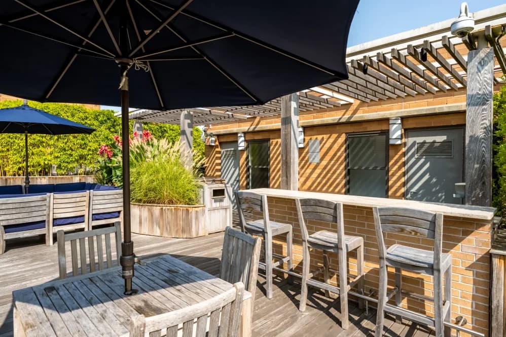 Rooftop terrace with wooden bar seating, black umbrellas, pergola, and brick building with green walls.