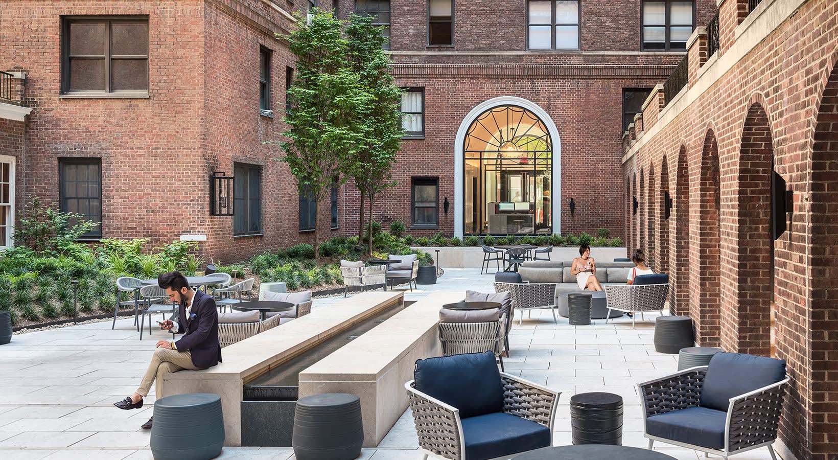 Brick courtyard with woven lounge chairs, built-in benches, potted plants, and ivy-covered building facade.