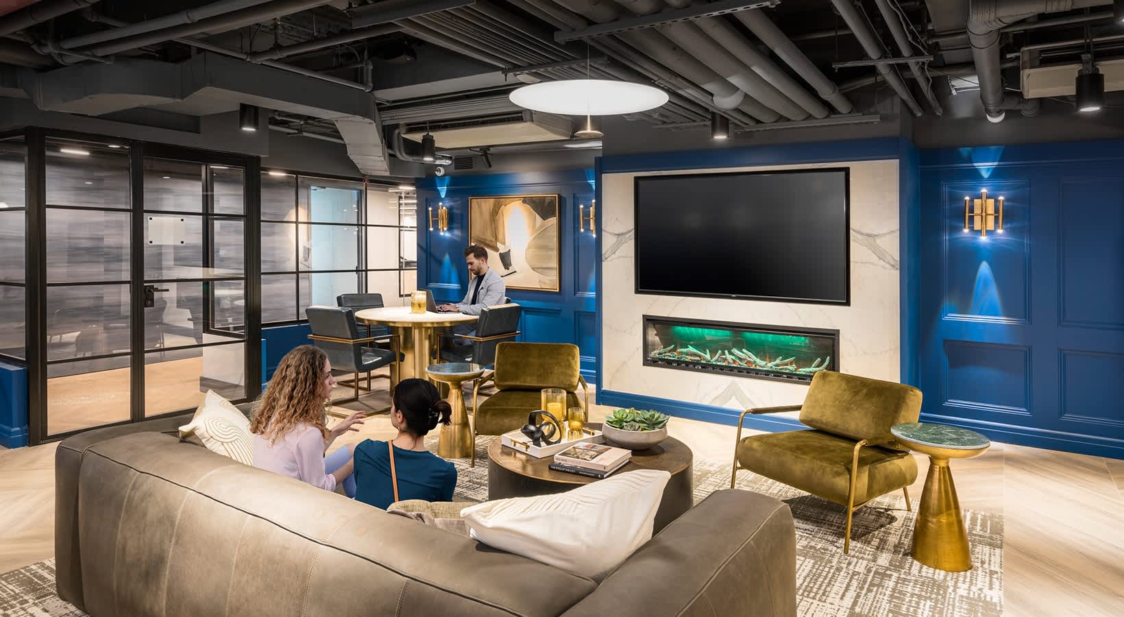 Open-concept lounge with gray sectional sofa, olive green armchairs, wall-mounted TV with fireplace, and blue accent walls with industrial ceiling.