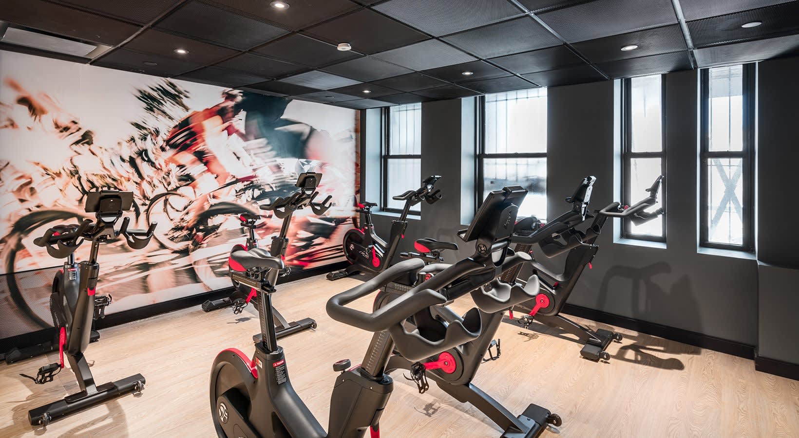 Fitness studio with multiple stationary bikes and exercise equipment, large windows, and red and black cycling mural on wall.