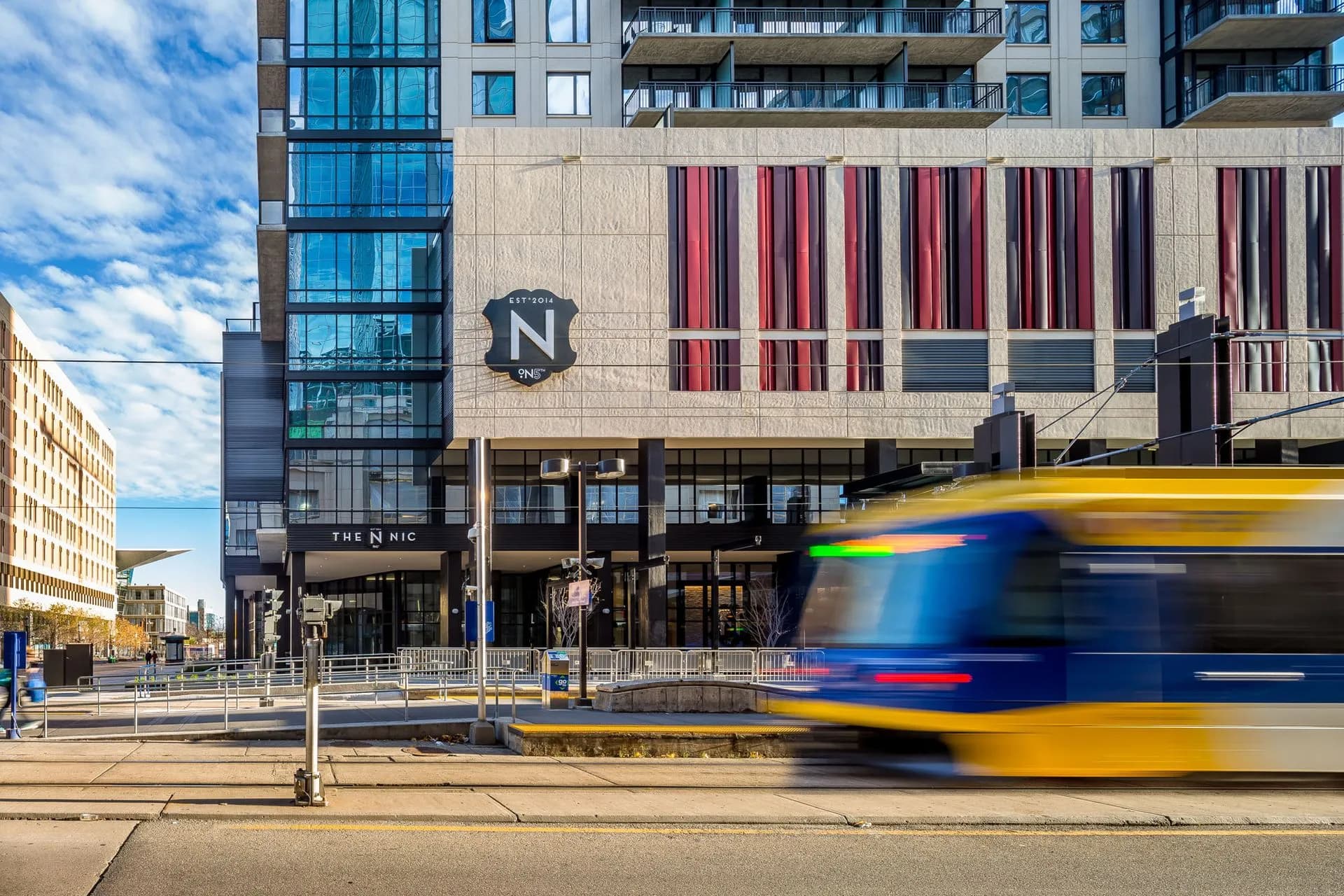 Modern multi-story residential building with glass balconies, striped facade panels, and ground-floor retail, with a blue and yellow transit bus passing in front.