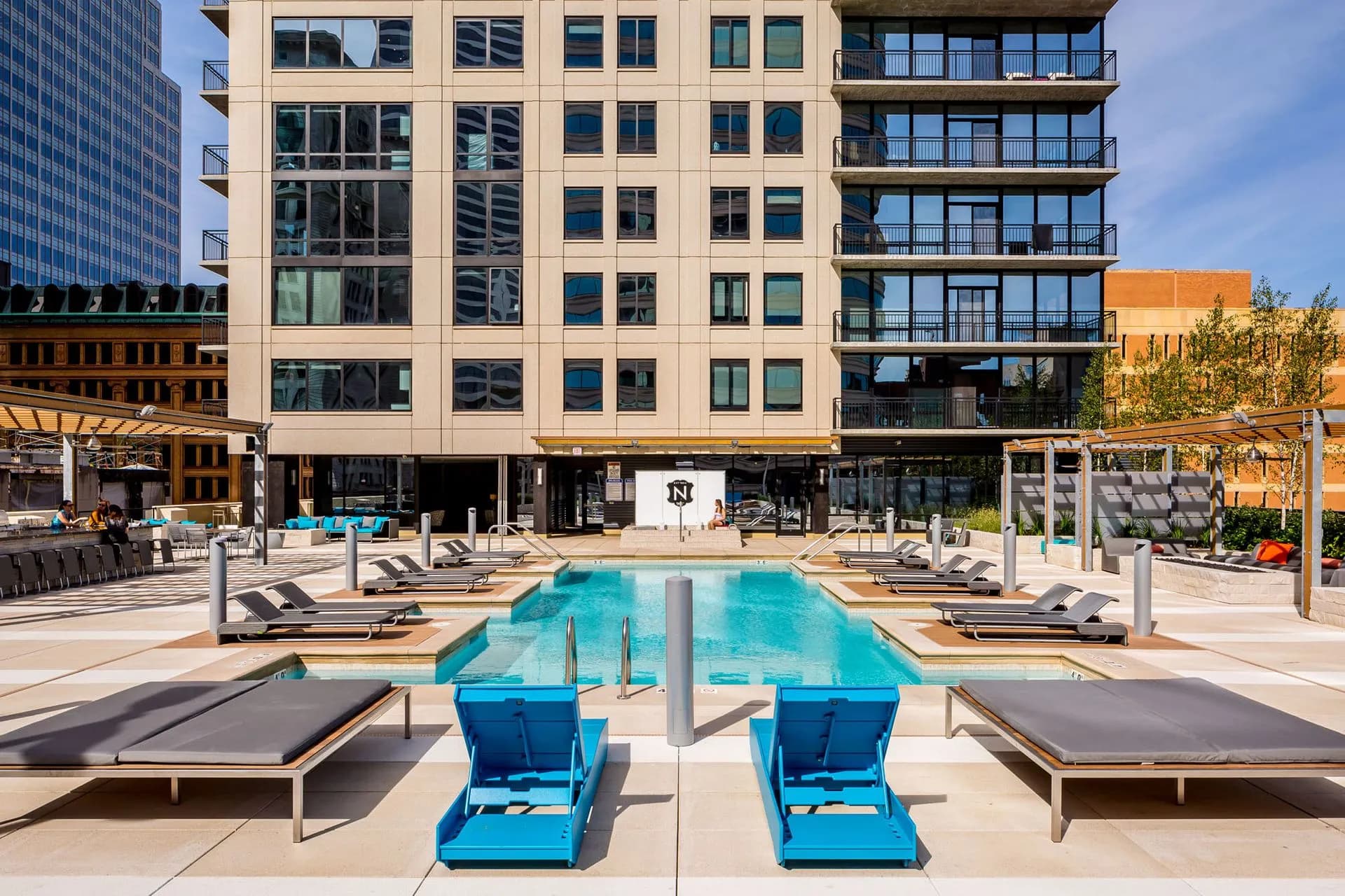 Resort-style pool with blue lounge chairs, concrete deck, and multi-story residential building with balconies in background.