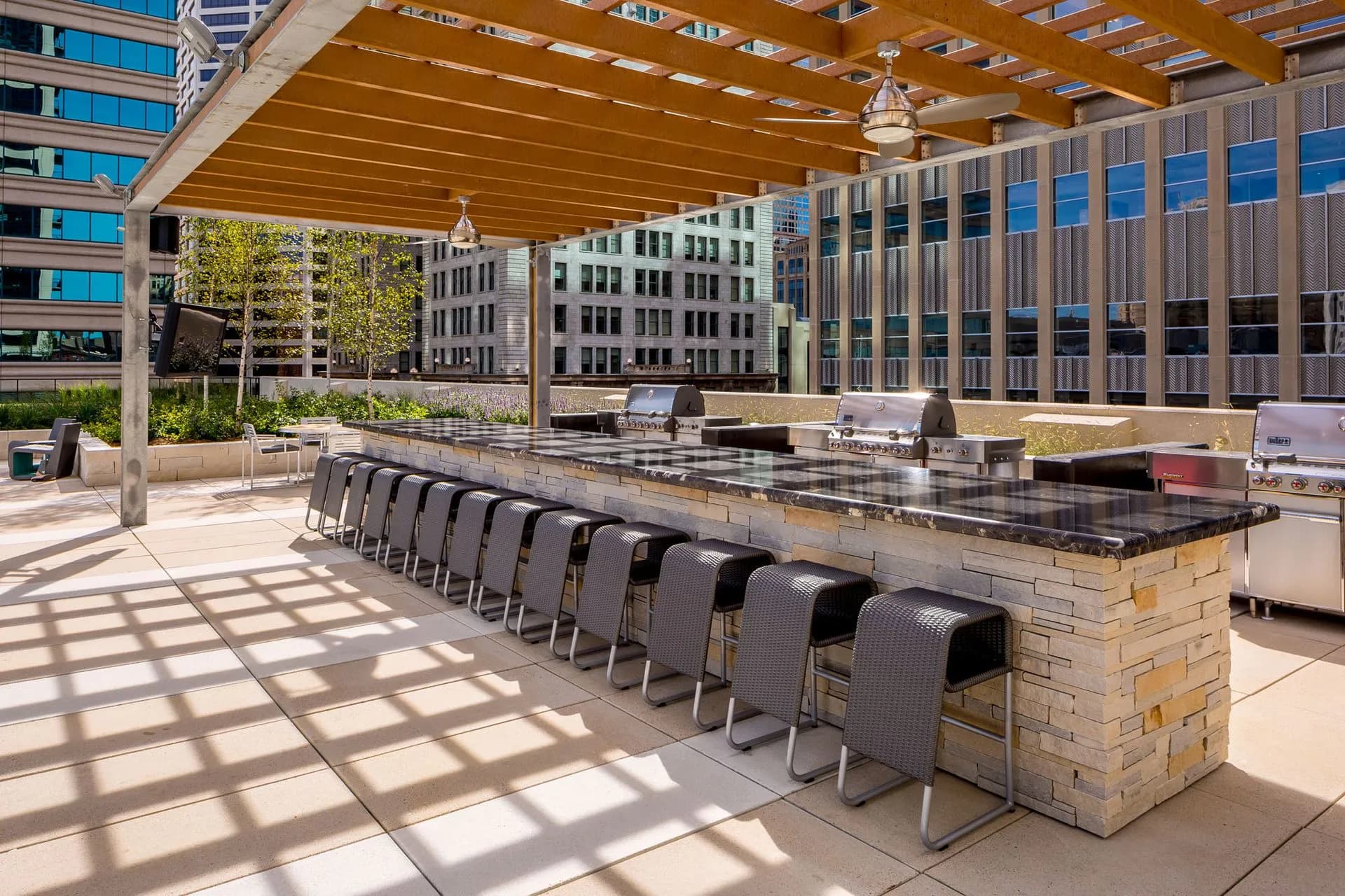 Rooftop terrace with wooden pergola, long stone counter with built-in grills, black dining chairs, and city views.