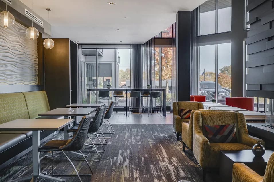 Contemporary lounge with mustard armchairs, dining tables, pendant lights, and expansive windows overlooking autumn trees.