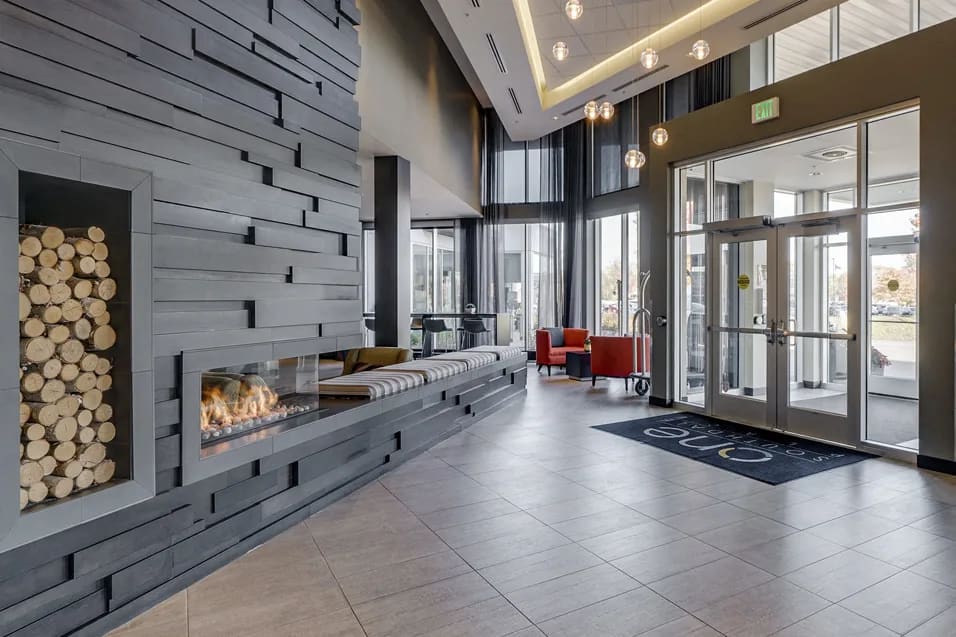 Contemporary lobby with horizontal fireplace, wood-slat accent wall, pendant lighting, and floor-to-ceiling glass doors opening to exterior.