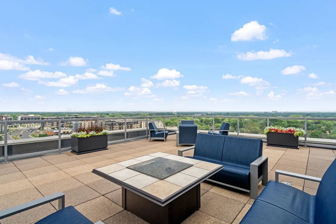 Rooftop terrace with blue seating, fire pit table, planters, and expansive views of Edina landscape under clear sky.