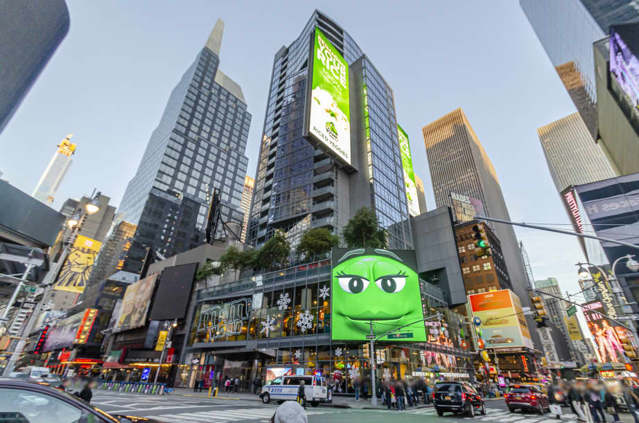 Street-level view of 1600 Broadway tower in Times Square with digital billboards, surrounding skyscrapers, and busy pedestrian intersection.