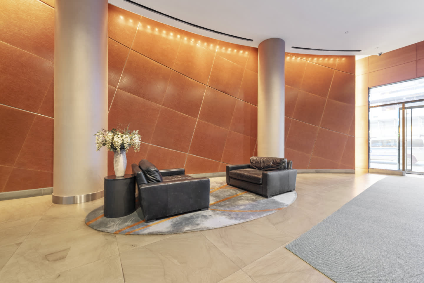 Building lobby with two black leather chairs on a patterned rug, cylindrical column, terracotta accent walls with ambient lighting, and marble flooring.