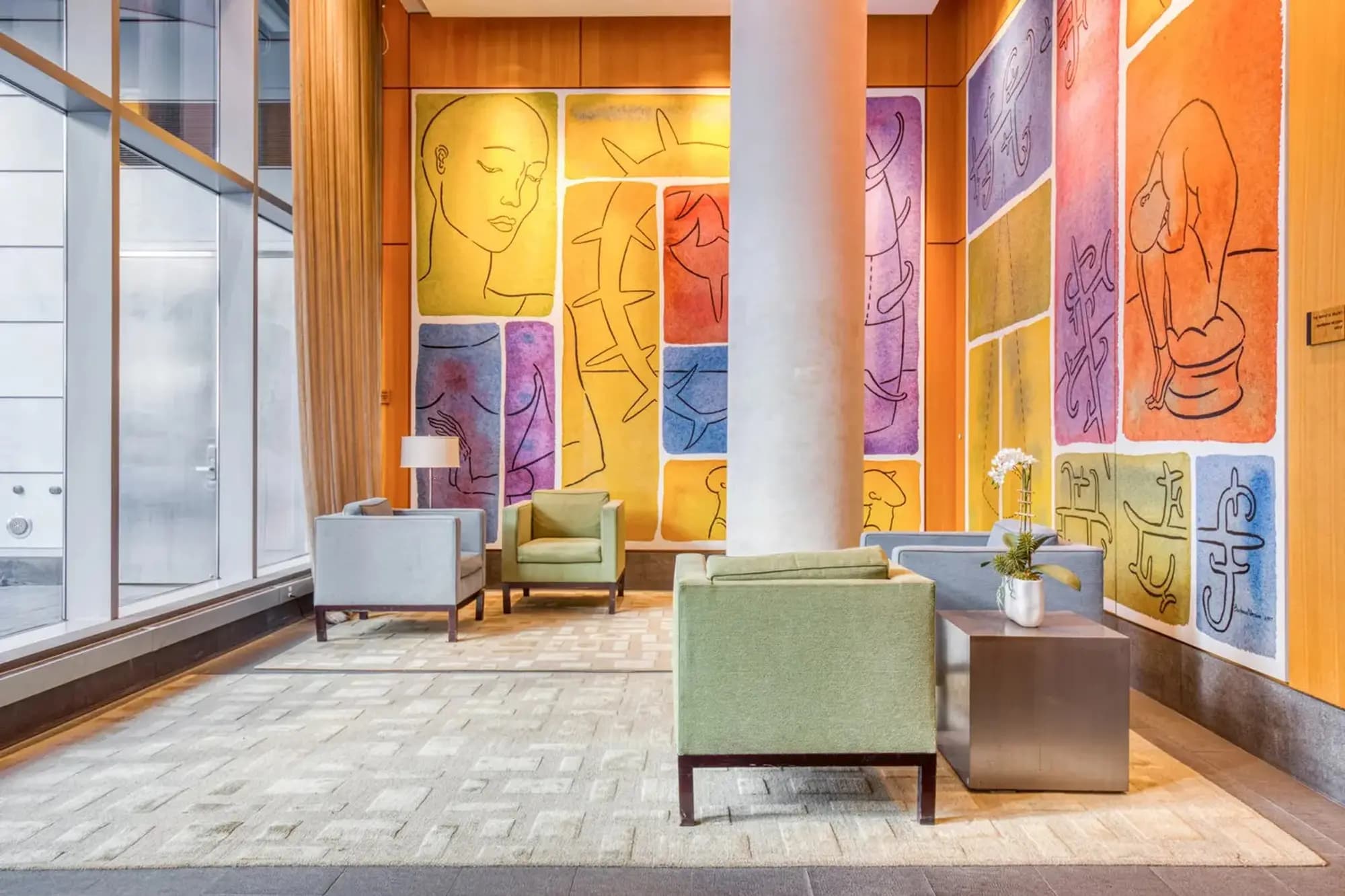 Modern lobby with green upholstered chairs, colorful abstract art panels in orange and purple tones, and large windows with city views.