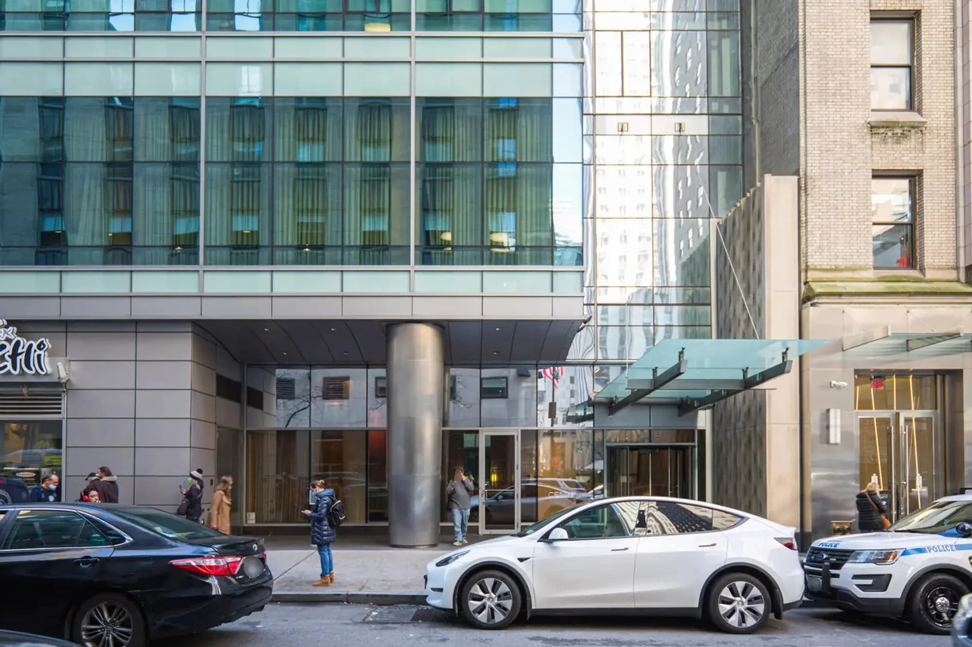 Modern glass and steel building facade with street-level entrance, parked vehicles, and pedestrians on urban sidewalk.
