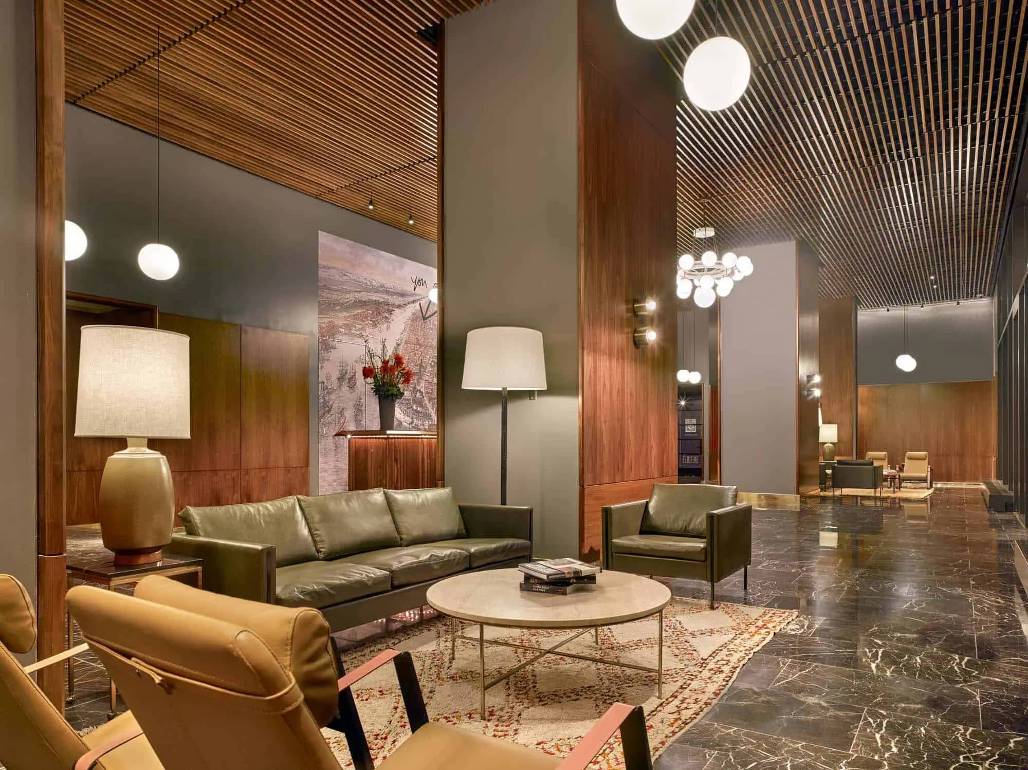 Modern lobby lounge with olive green sofa, tan armchairs, round marble table, wood-slat ceiling, and recessed lighting.