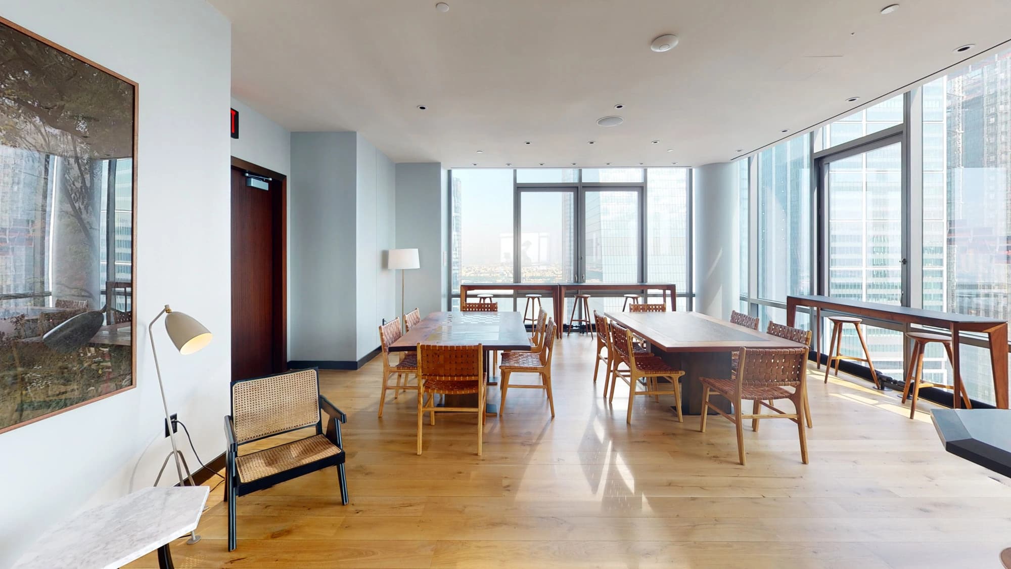 Open common area with wooden dining tables, mid-century chairs, hardwood floors, and expansive windows with city views.