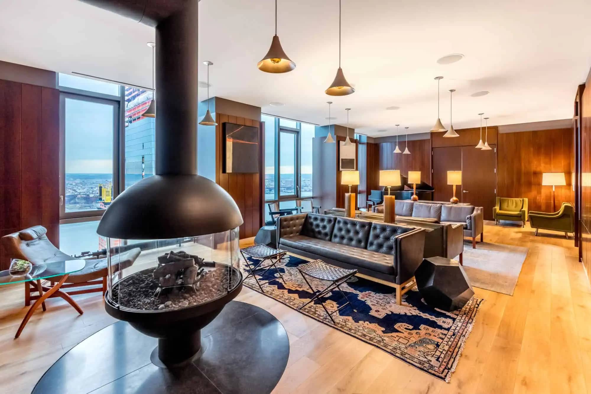 Modern living room with black spherical fireplace, leather sofa, pendant lights, patterned rug, and floor-to-ceiling windows overlooking the city.