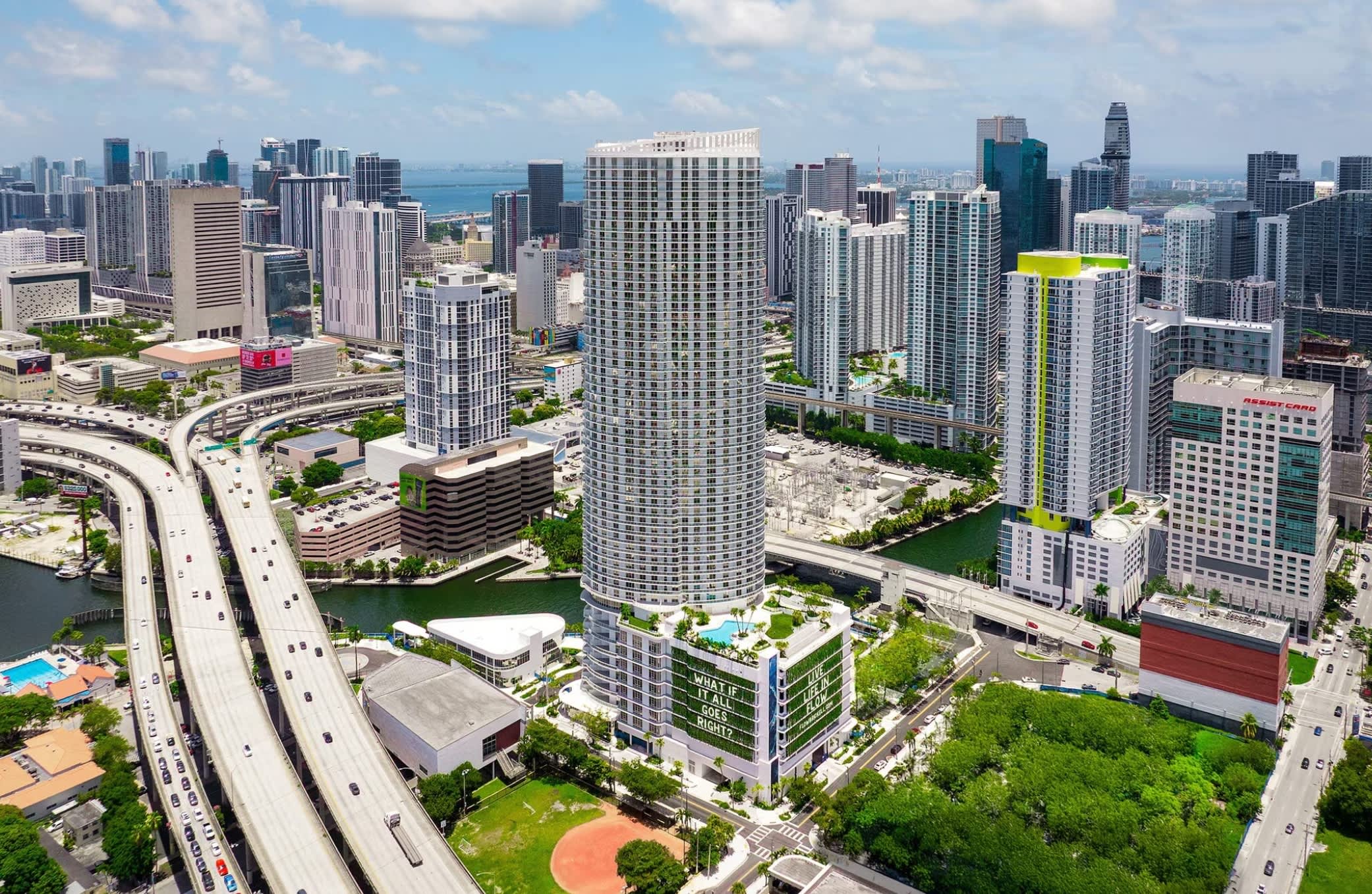 Aerial view of Flow Brickell, a tall residential tower in downtown Miami surrounded by other high-rises, green spaces, and the Chicago River.