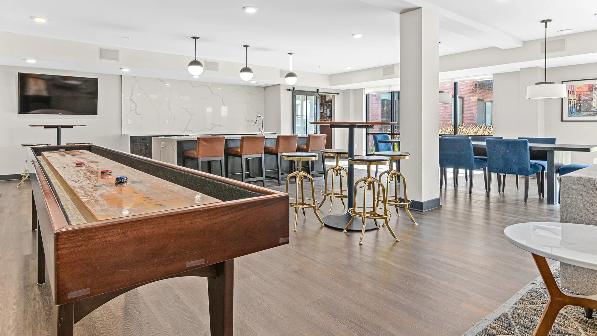Open common room with wooden shuffleboard table, bar seating with brown and blue chairs, kitchen, and lounge area with pendant lighting.