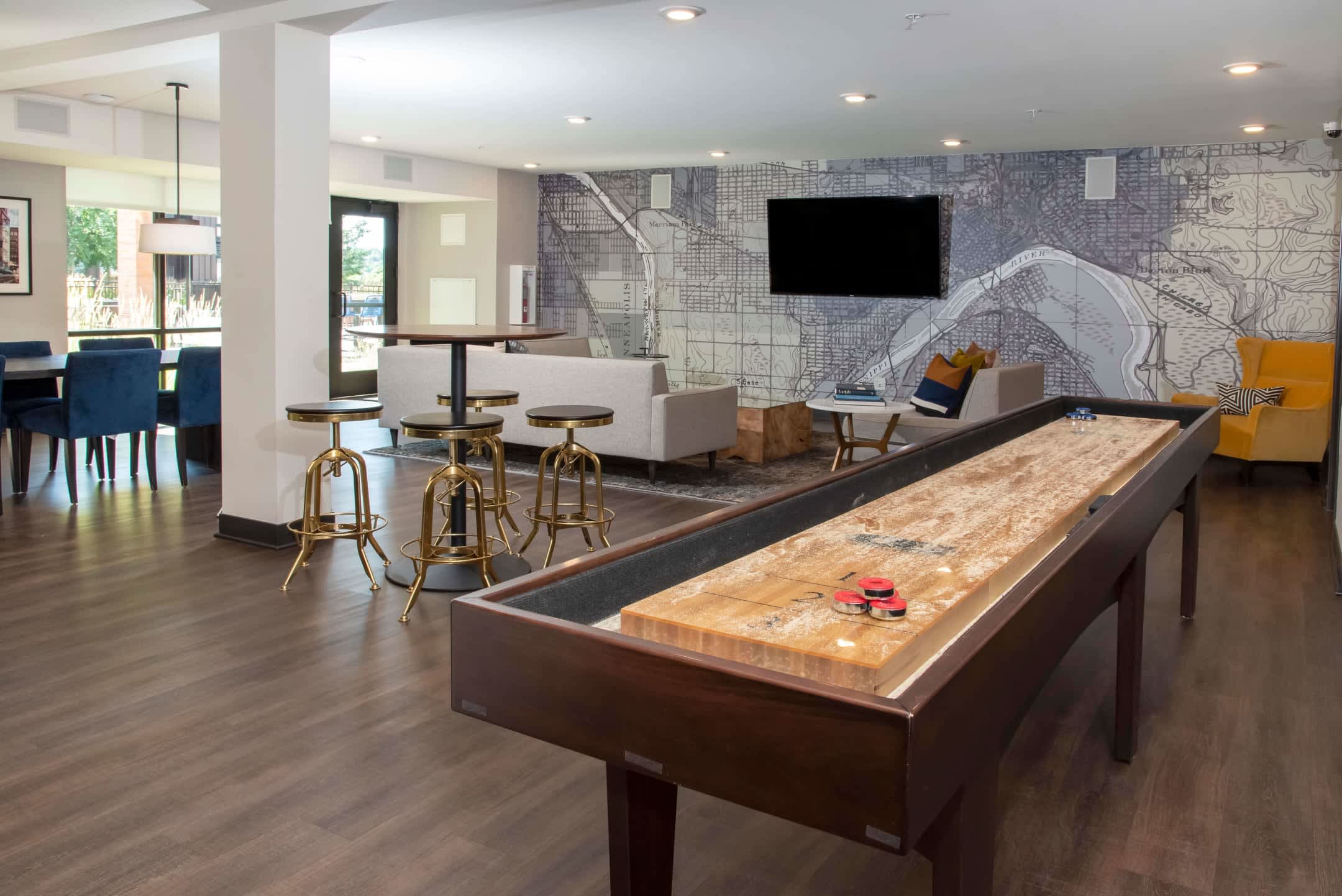 Common room with shuffleboard table in foreground, bar seating with brass stools, lounge area with gray sofa, wall-mounted TV, and map-patterned accent wall.