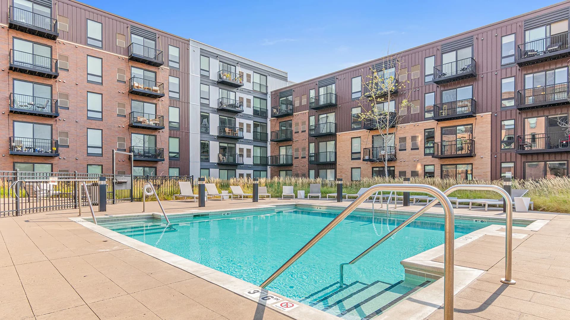 Outdoor swimming pool with metal handrails and entry steps, surrounded by tan concrete deck, lounge chairs, and multi-story brick apartment buildings under clear blue sky.