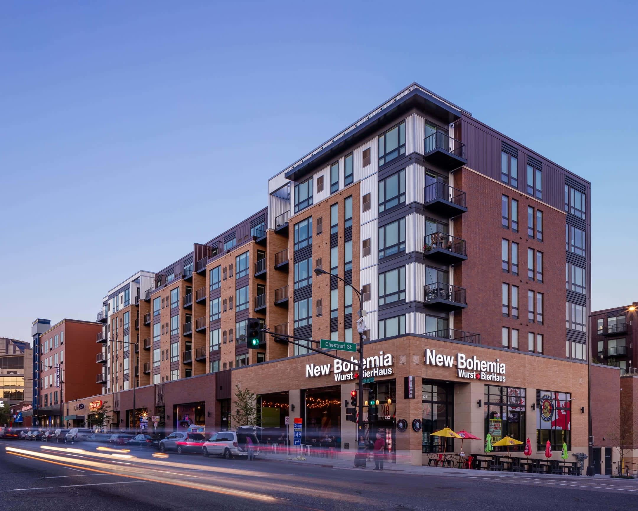 Multi-story brick apartment building with retail storefronts at street level, balconies on upper floors, and clear evening sky.