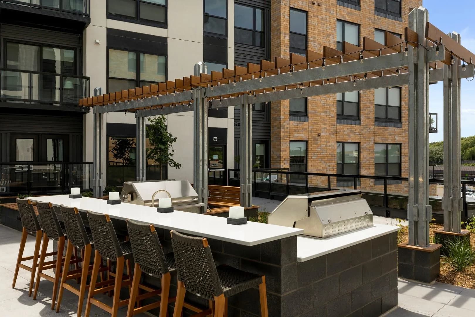 Rooftop terrace with white countertop bar, woven bar stools, stainless steel grill, and wooden pergola overlooking brick residential buildings.