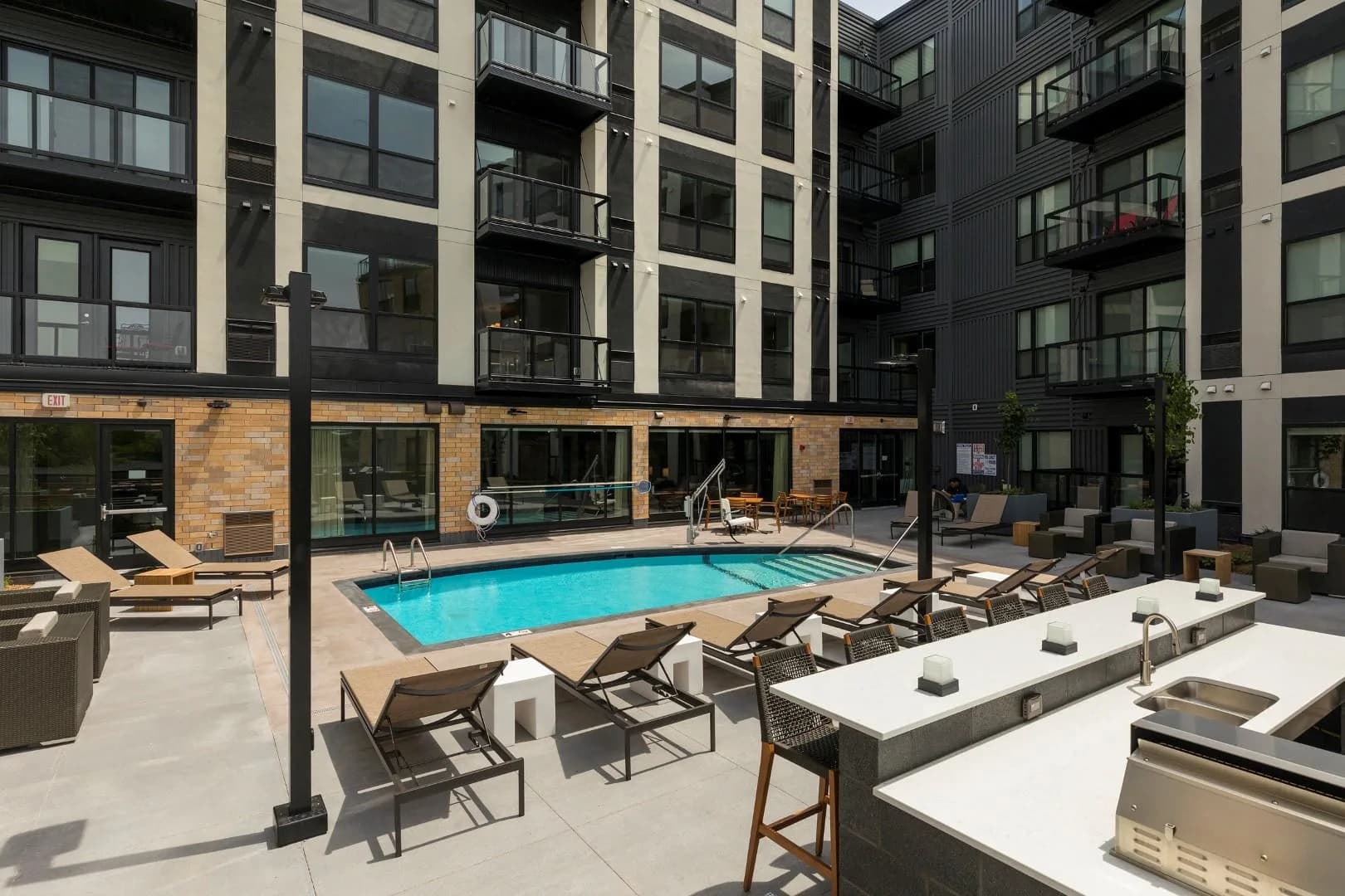 Courtyard with bright blue swimming pool, lounge chairs, white dining tables with grills, and multi-story residential buildings with brick and metal facades.