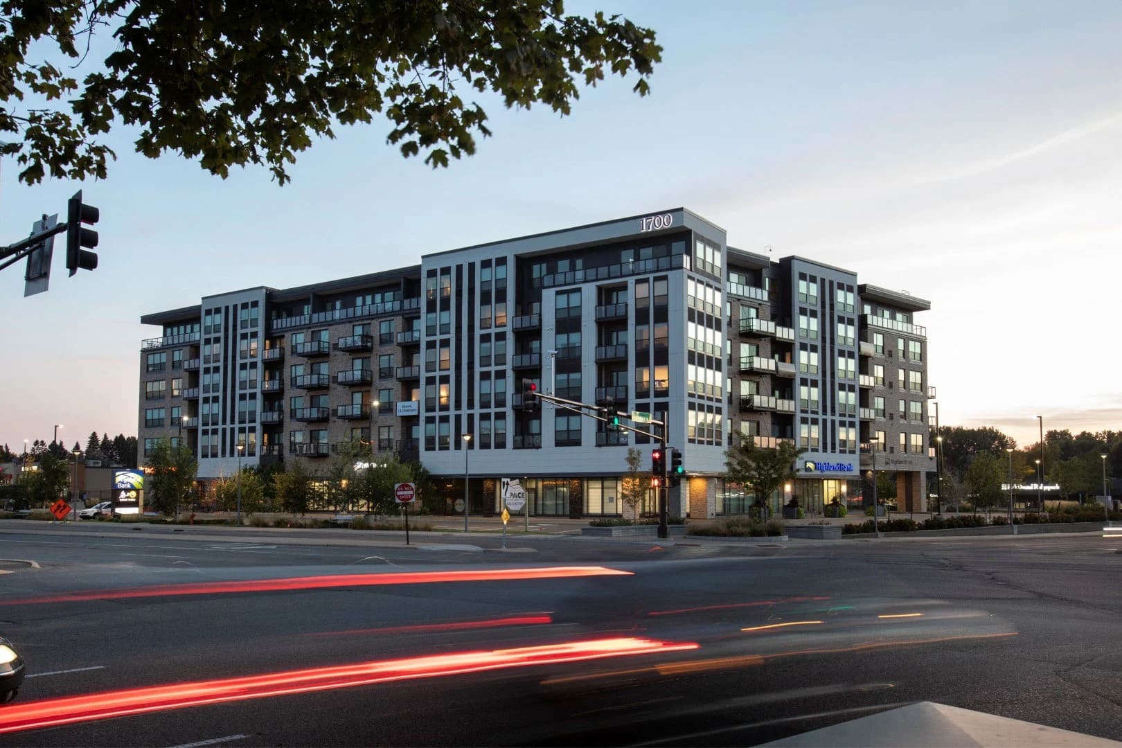 Modern six-story residential building with blue-gray facade, ground-floor retail spaces, and tree-lined street frontage at dusk.