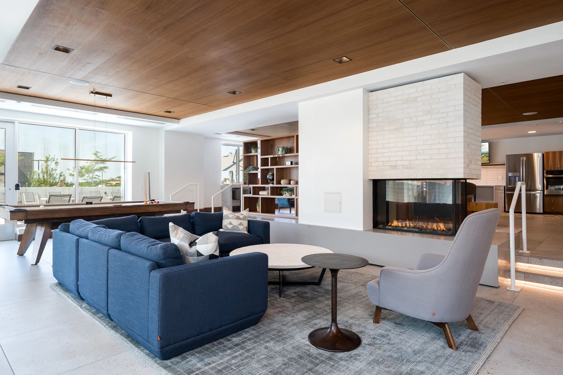 Modern living room with blue sectional sofa, fireplace with white surround, wood ceiling, and built-in shelving.