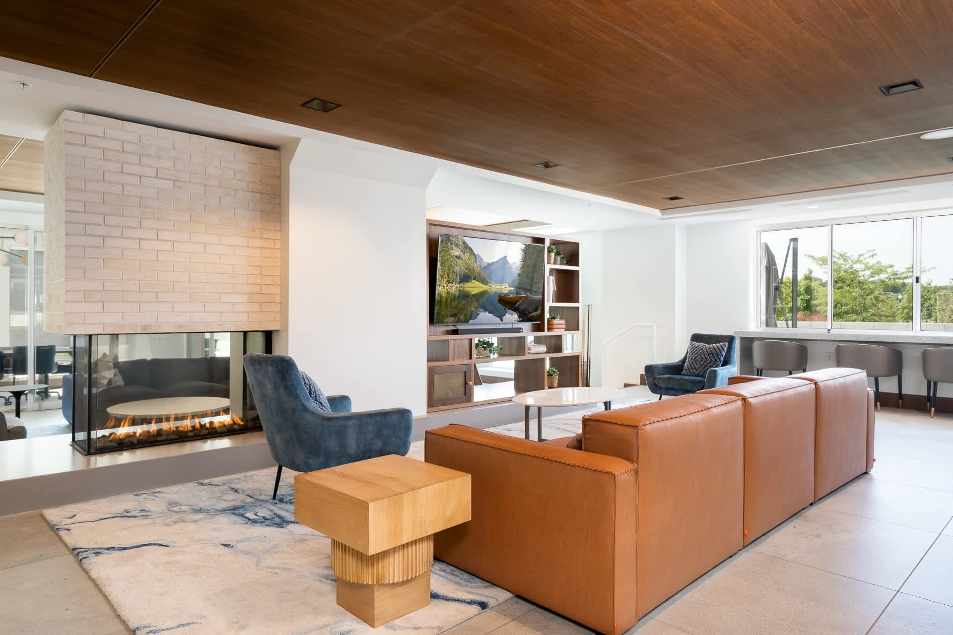 Modern lounge with orange leather sectional, blue accent chair, wood coffee table, brick fireplace, and large windows overlooking greenery.