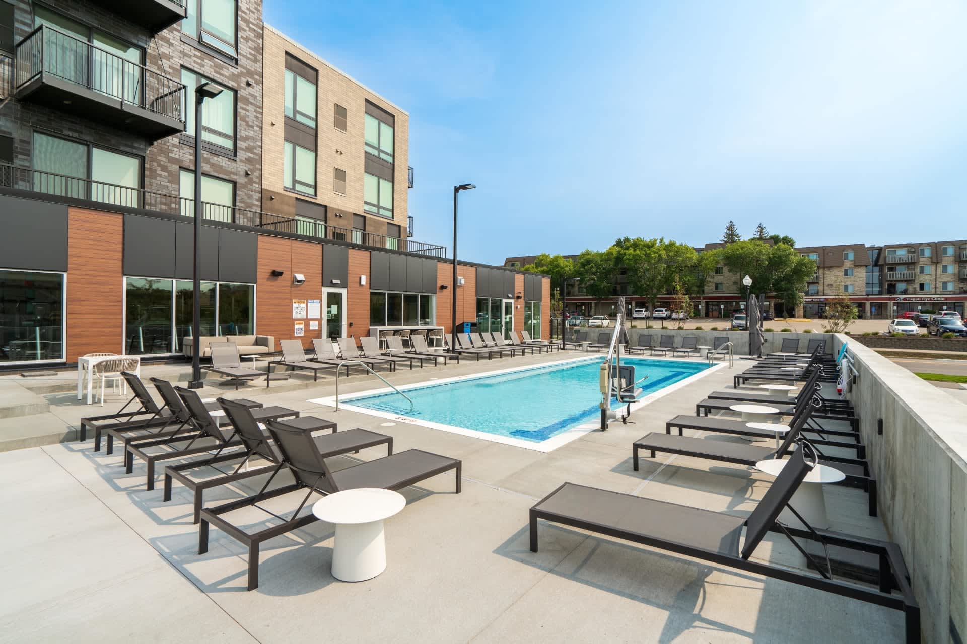 Outdoor pool deck with blue swimming pool, black lounge chairs, white umbrellas, and modern brick apartment building under clear sky.