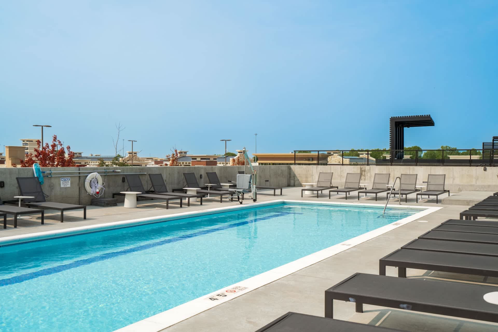 Rooftop swimming pool with blue water, concrete deck, black lounge chairs, and gray seating area under clear sky.