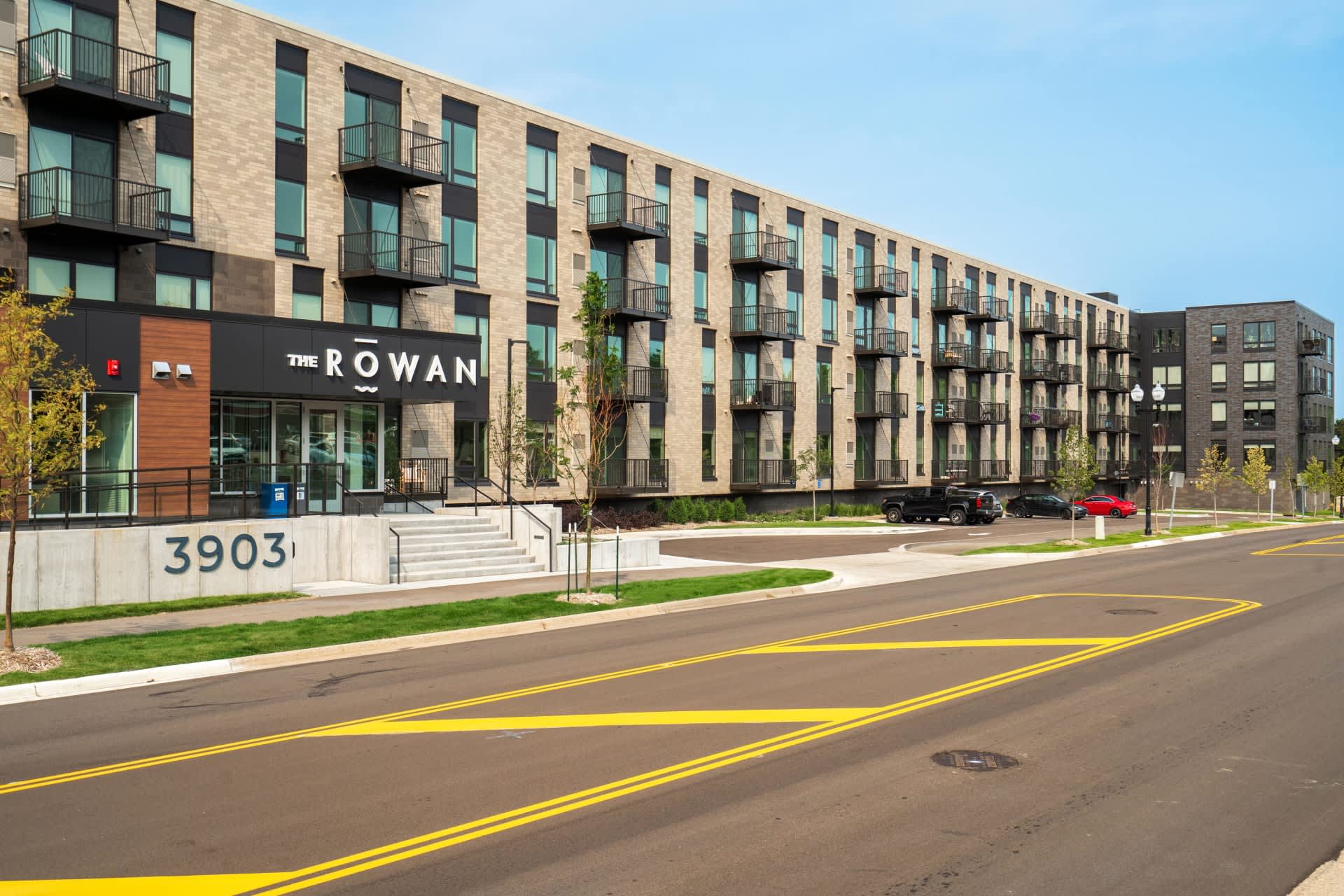Modern multi-story apartment building with brick and dark facade, ground-floor retail storefront labeled The Rowan, black metal balconies, and street-level parking.