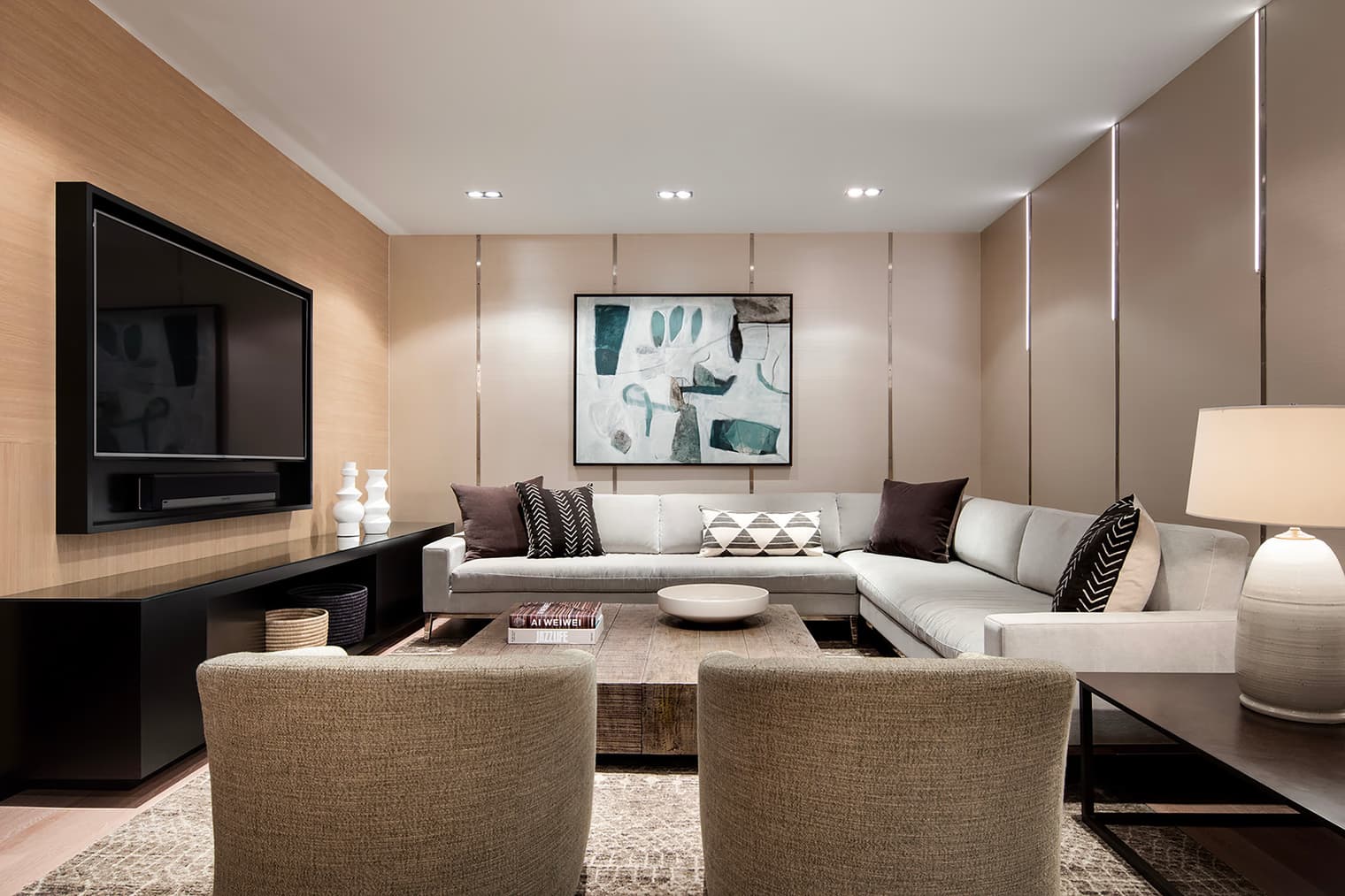Modern living room with cream sectional sofa, two tan armchairs, wall-mounted flat-screen TV, and abstract wall art.