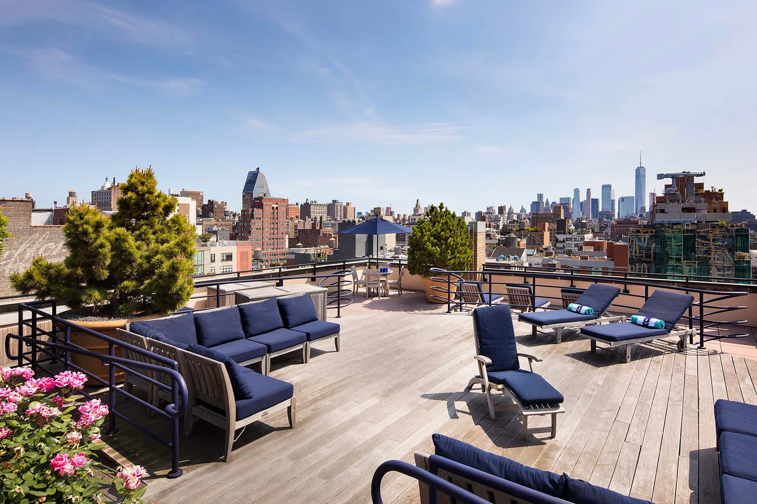 Rooftop terrace with blue cushioned seating, potted evergreens, pink flowers, and panoramic Manhattan skyline view.